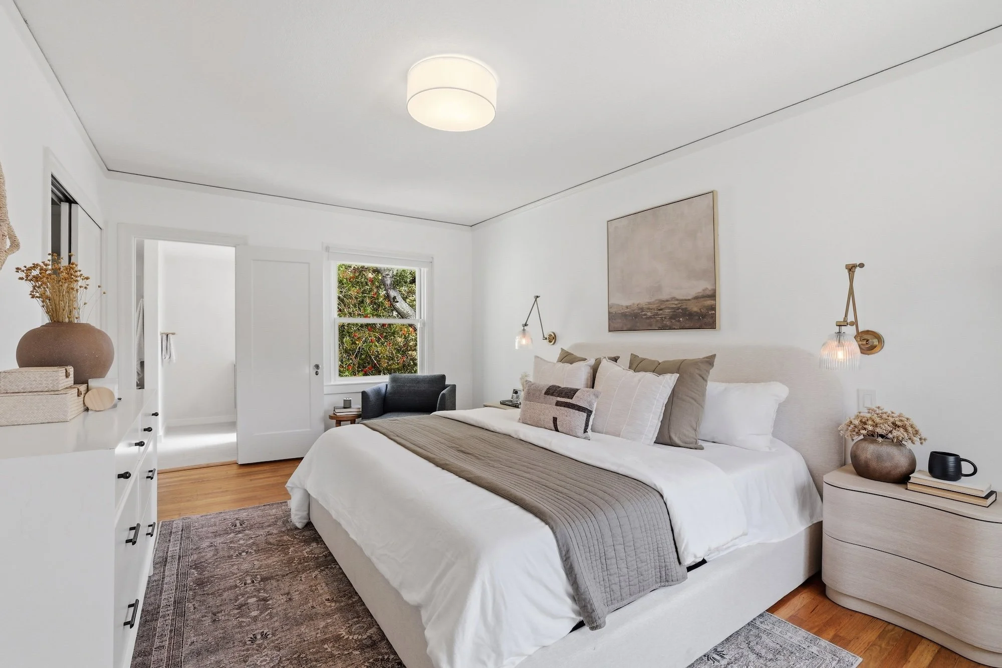 A bedroom with white walls and a white ceiling, featuring a bed with beige pillows and a gray blanket. There are two wall-mounted lamps beside the bed, a painting above, a window showing trees outside, a nightstand with a vase and books, and a dresse