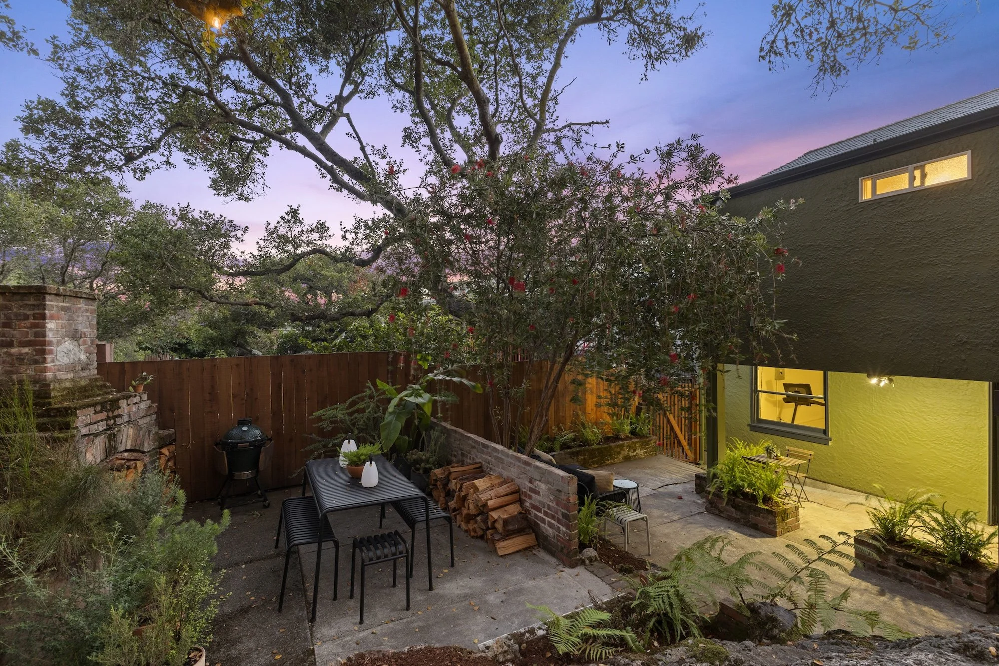 A backyard garden area with a large tree, a brick wall, a wooden fence, a small patio with outdoor furniture, and a yellow house with a window. The sky shows a colorful sunset.