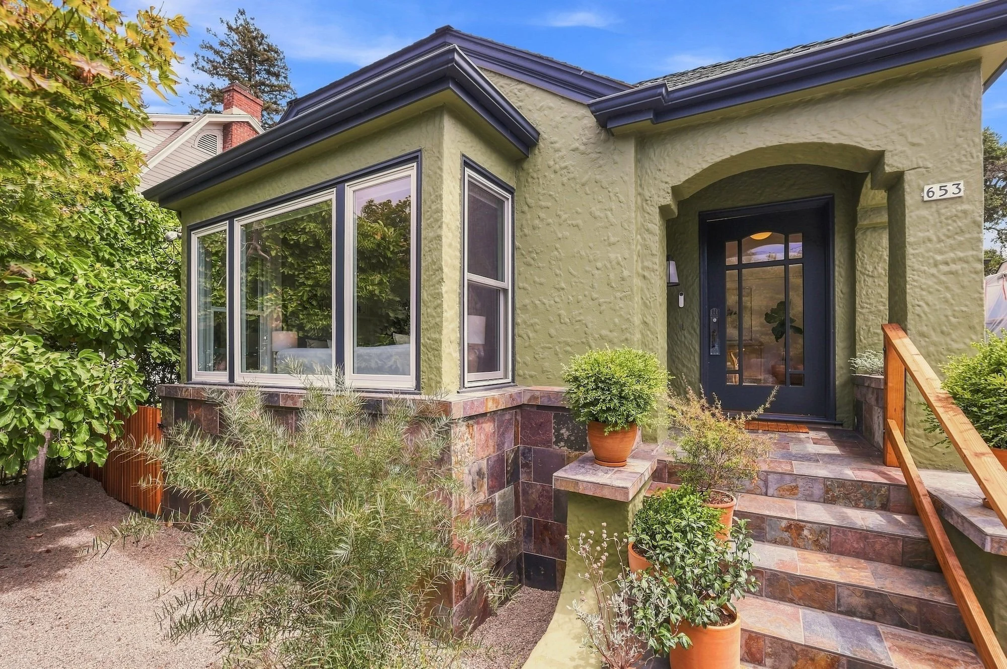 Front porch of a house with a black front door, large window, potted plants, stone steps, and a garden with shrubs and trees.