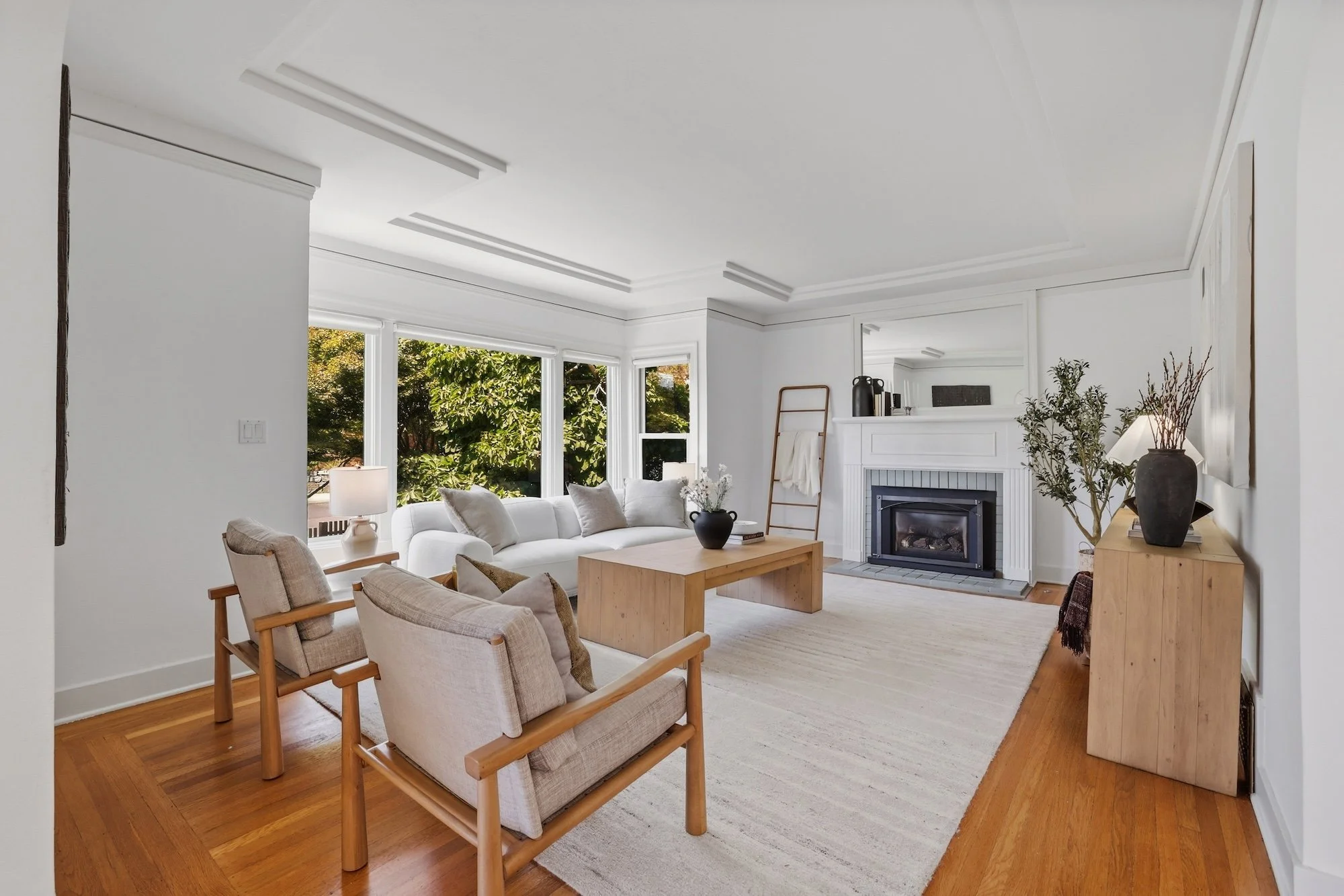 A bright living room with white walls, hardwood floors, large bay windows revealing greenery outside, featuring beige and white furniture, a black fireplace with a mantle, and various decorative plants and vases.