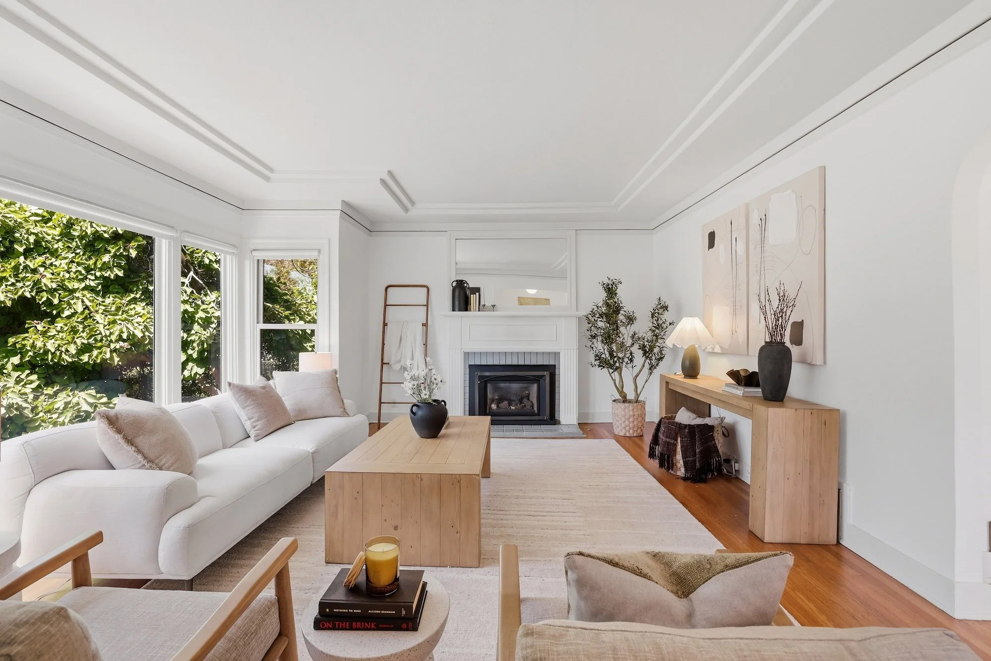 Cozy living room with white sofa, wooden coffee table, fireplace, and large windows with lush greenery outside. Decor includes abstract wall art, plants, and lamps.