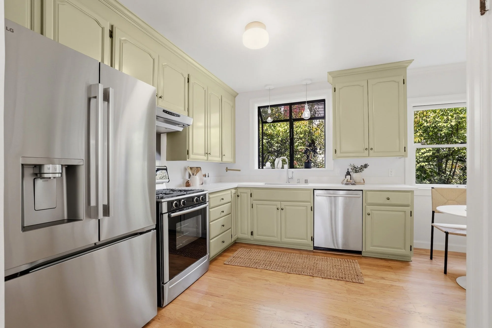 A kitchen with stainless steel appliances, beige cabinets, a white countertop, and two large windows showing green trees outside.