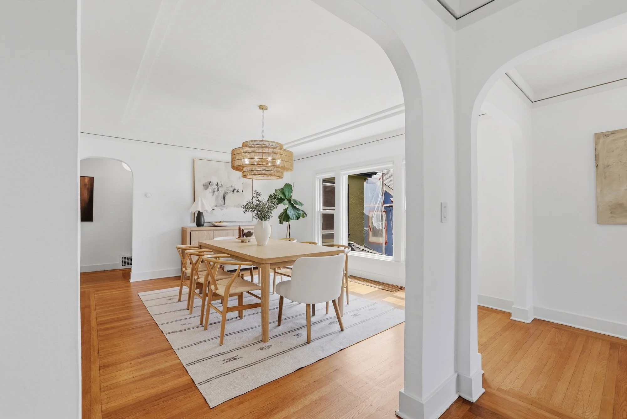 A dining room with a wooden table, beige chairs, a large window, a brass chandelier, white walls, and hardwood flooring.