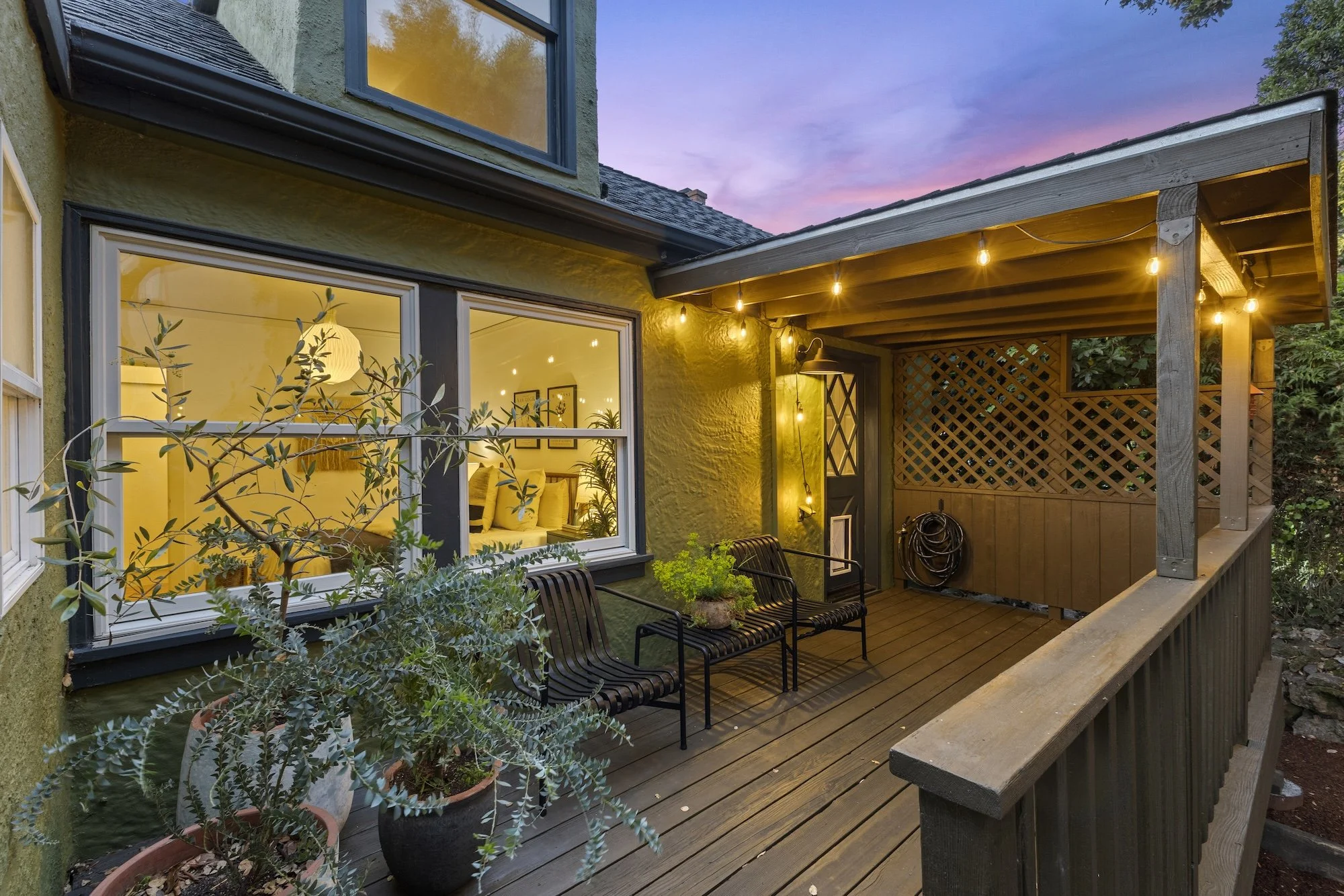 View of a cozy outdoor patio area at dusk with string lights, potted plants, and a view through the windows into a well-lit indoor room with comfortable furniture.
