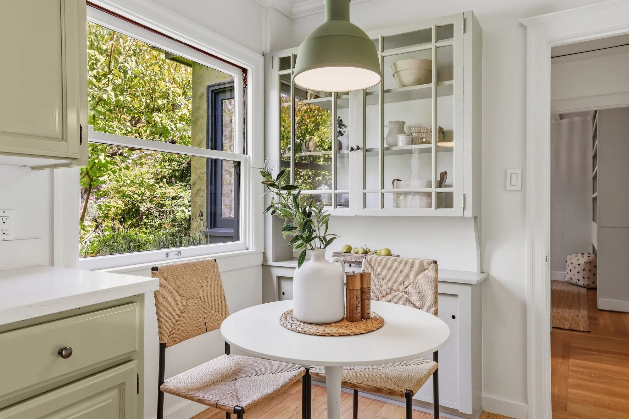 Kitchen with a round white table and three beige chairs, a white vase with a plant, and a window showing greenery outside.