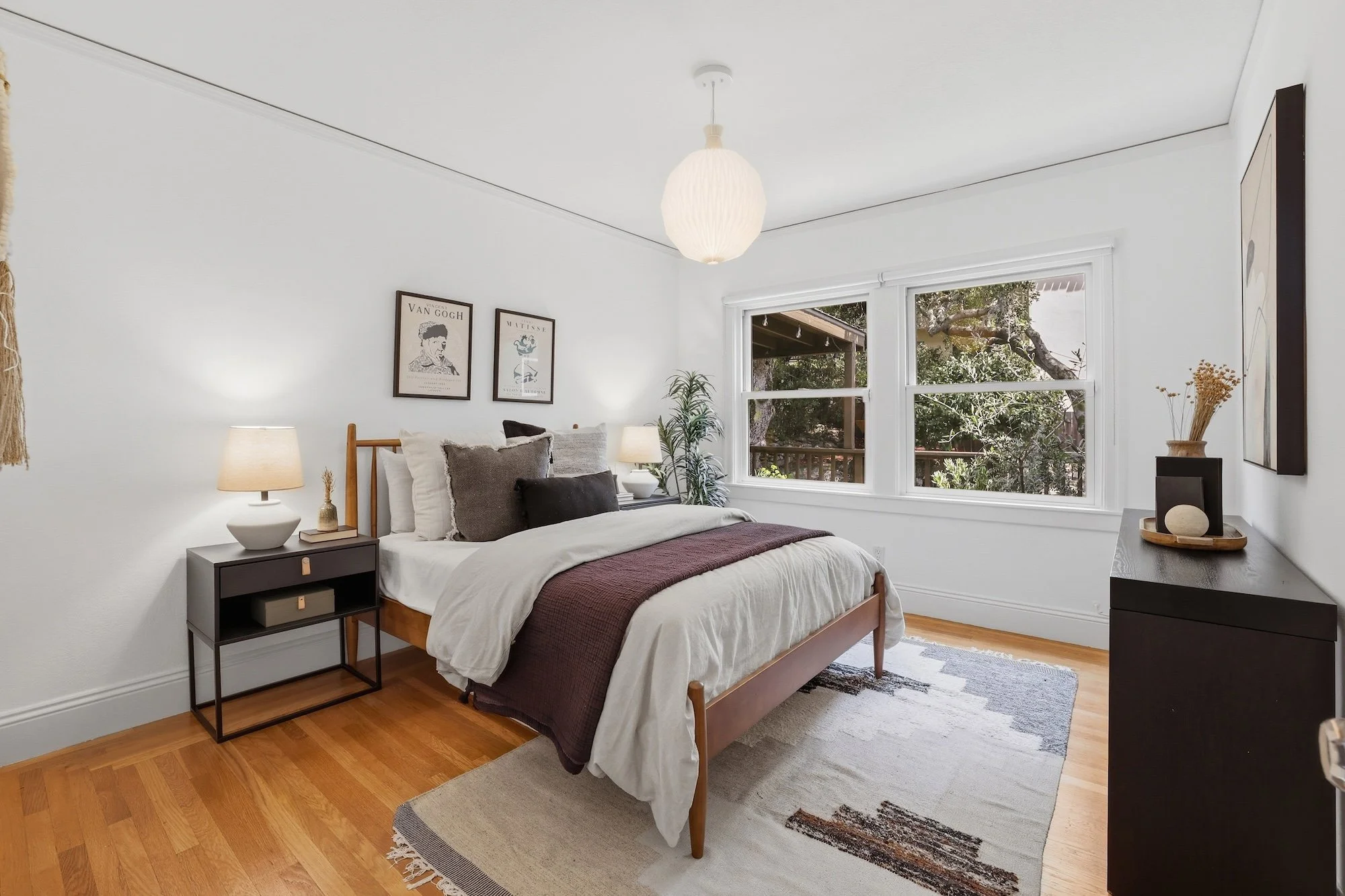 A bedroom with a wooden bed frame and white bedding, accented with dark gray and black pillows. Next to the bed is a black nightstand with a lamp, a decorative object, and a small plant. There is a window showing trees outside, and the walls are deco
