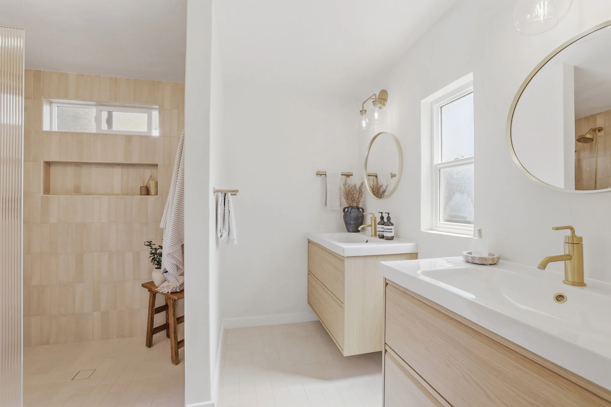 Modern bathroom with white walls, two sinks, light wood cabinets, and a mirror above each sink. There are two windows, and a shower area with beige tiles and a showerhead. Decorative items and toiletries are on the sink counter.