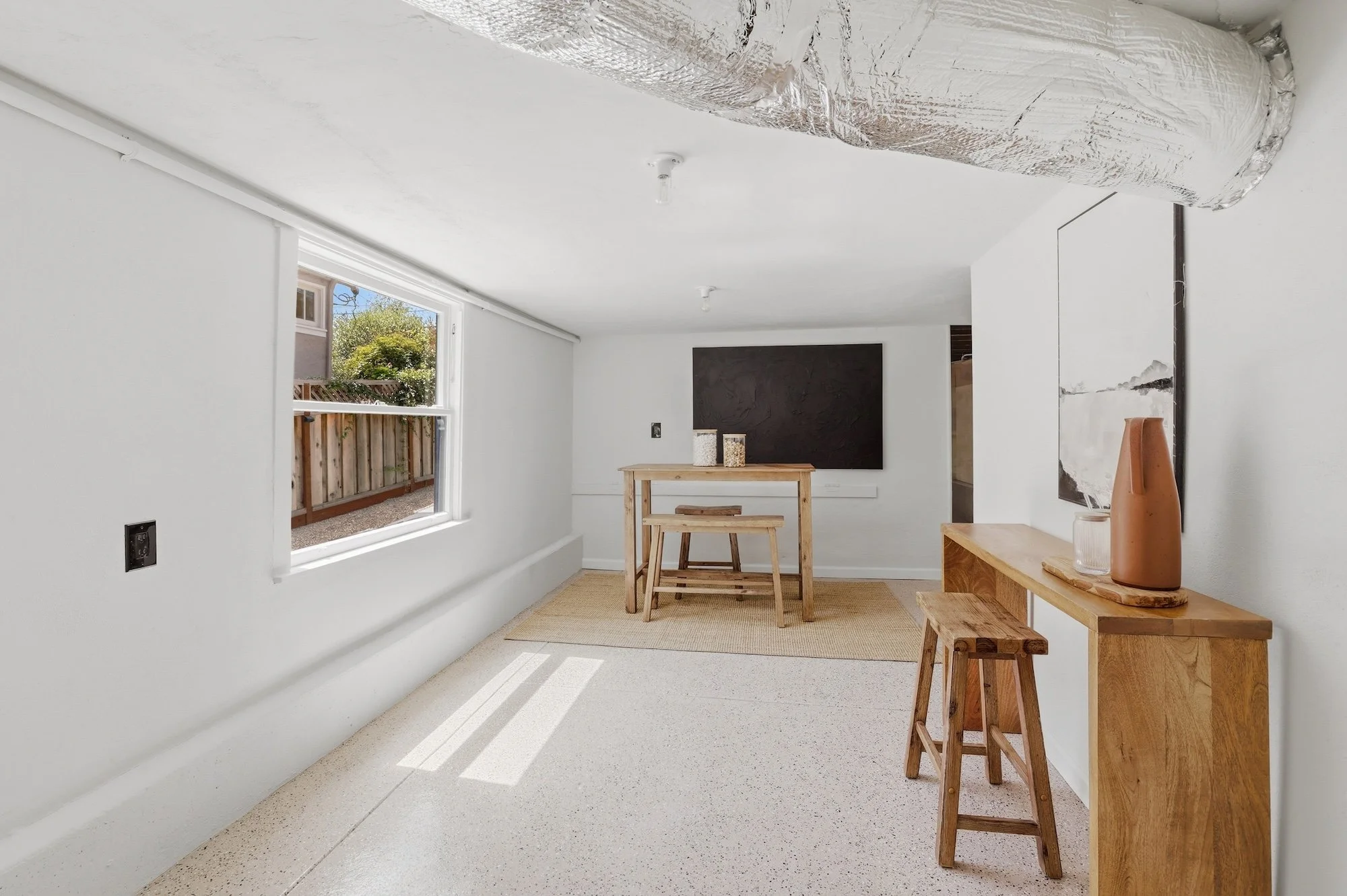 Interior of a room with white walls, window letting in natural light, and wooden furniture including a table and stools. The ceiling shows insulation material, indicating ongoing construction or renovation.