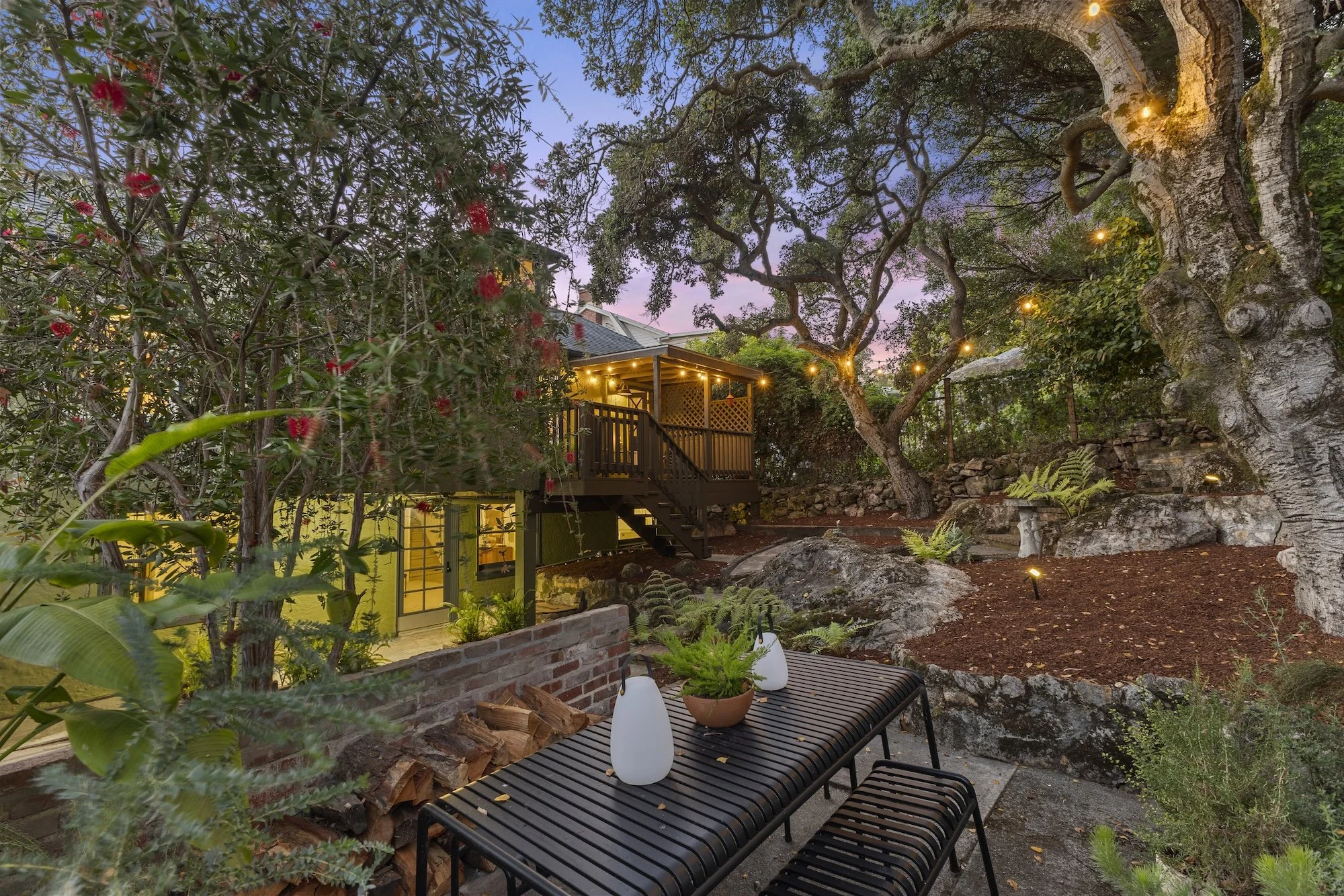 A backyard scene at dusk with a tree decorated with string lights, a wooden deck with outdoor furniture, and various plants and rocks. The sky has a pinkish-purple hue.