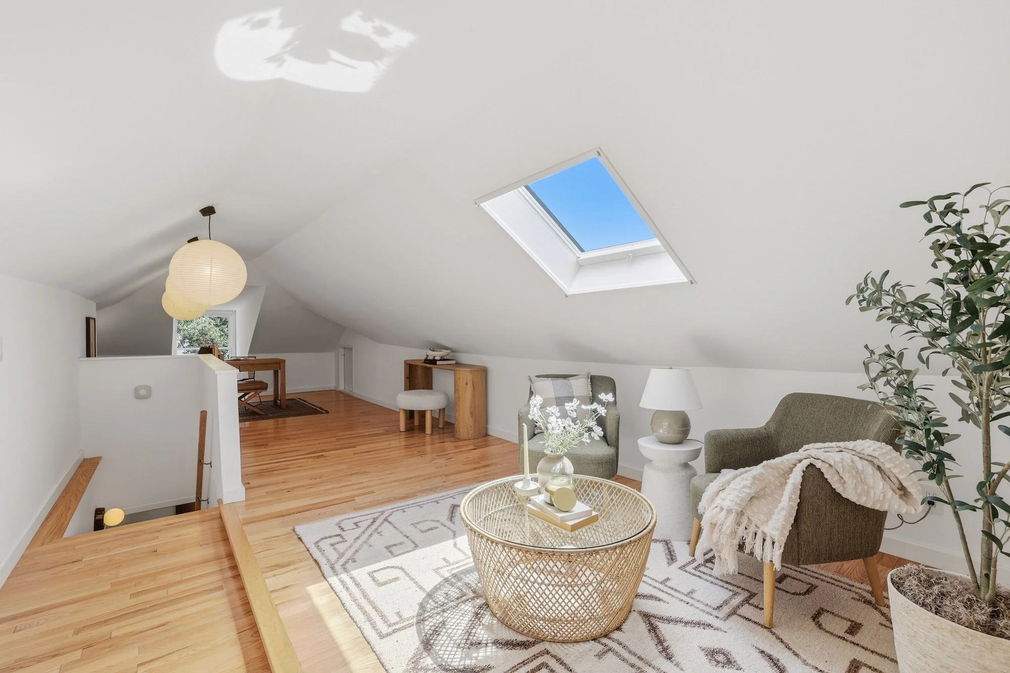 Living room with sloped white ceiling, skylight, wooden flooring, green armchair with blanket, white side table with lamp, decorative items, and houseplant.