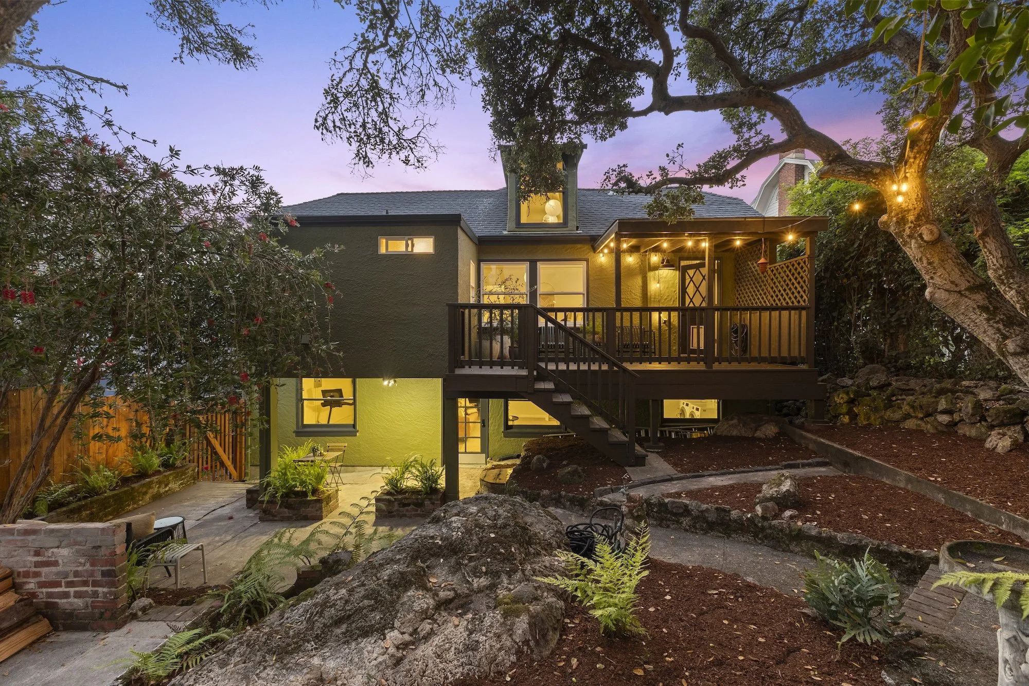 Backyard view of a two-story house with a green exterior, a wooden deck with string lights, and a surrounding garden with large rocks, plants, and trees at dusk.