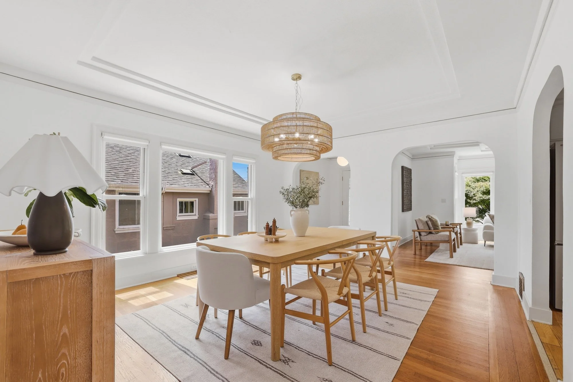 Bright, minimalist dining room with wooden table, surrounding chairs, large window, and modern light fixture.