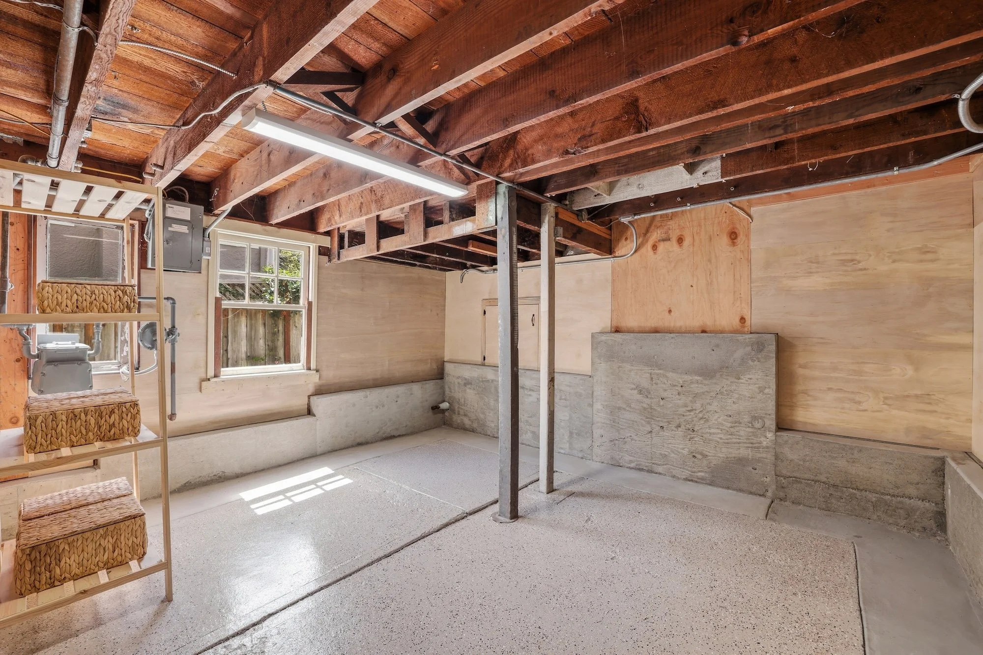 Interior of a room under construction with exposed wooden ceiling beams, a fluorescent light fixture, and a window allowing natural light. There are some concrete and wooden wall panels and unfinished flooring.
