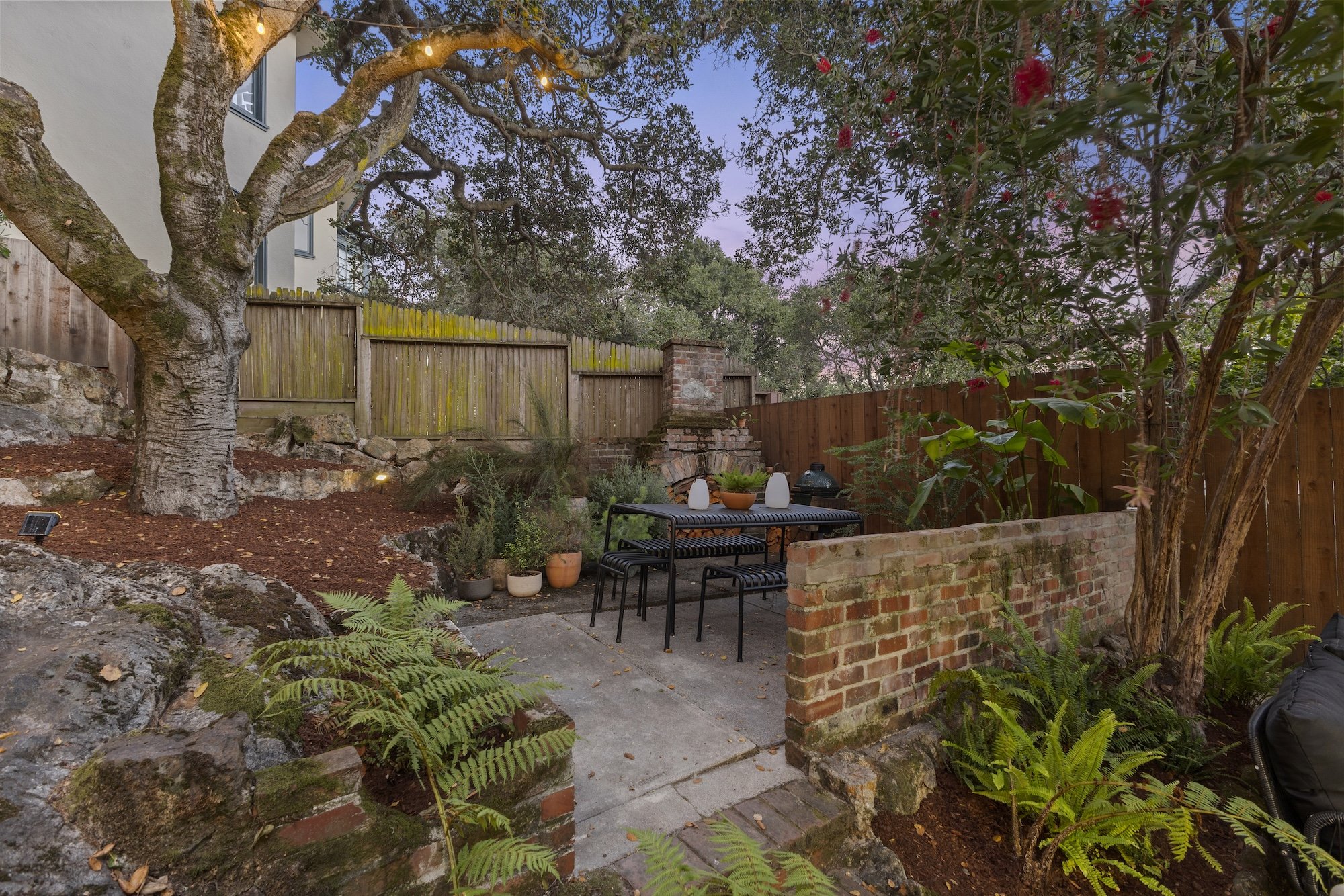 A backyard garden with trees, potted plants, a brick wall, and outdoor seating, taken during the early evening.