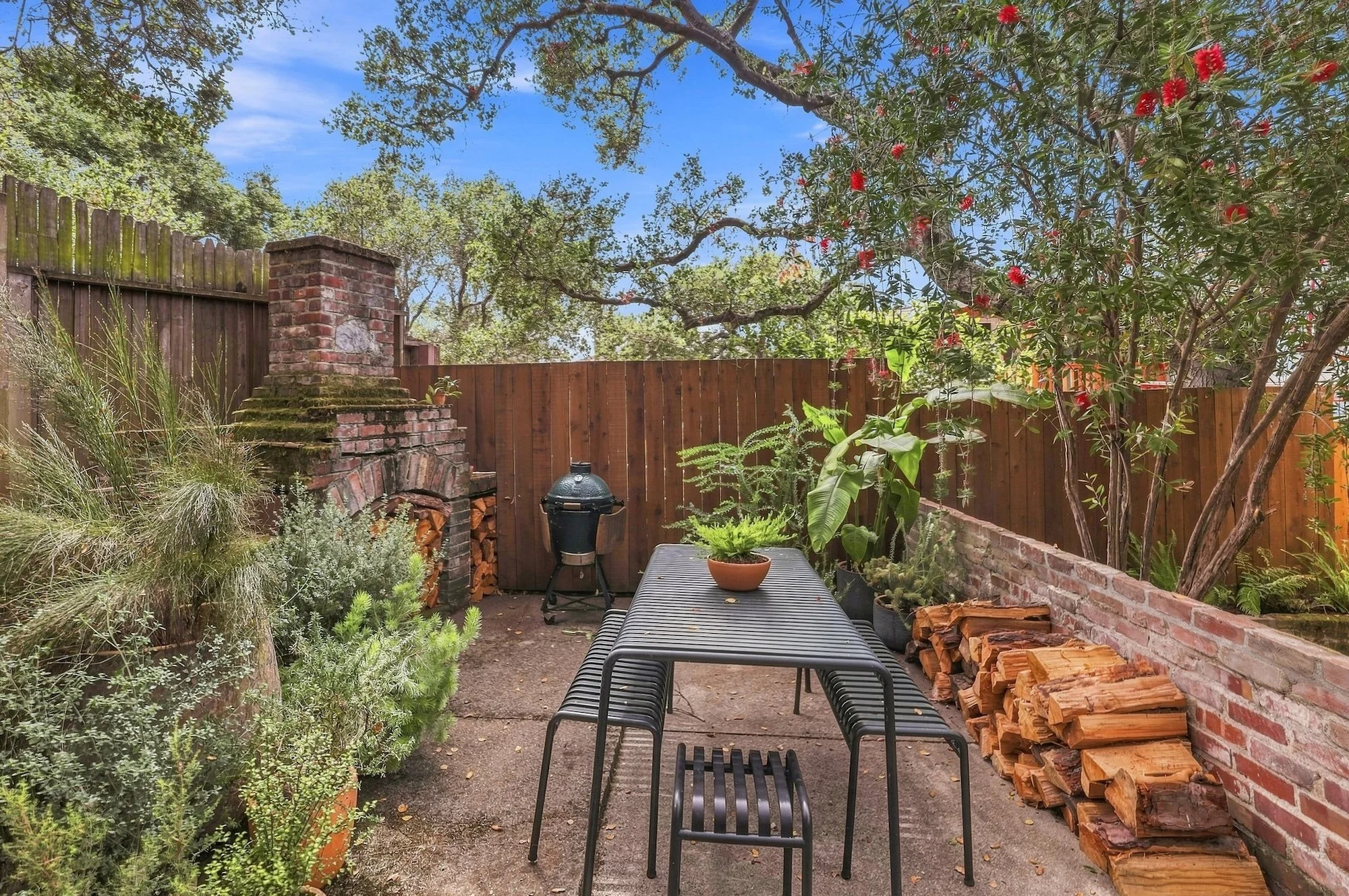 A backyard patio with a black metal table and benches, potted plants, a brick fireplace with firewood, and a wooden fence, surrounded by trees with green leaves and red flowers, under a blue sky.