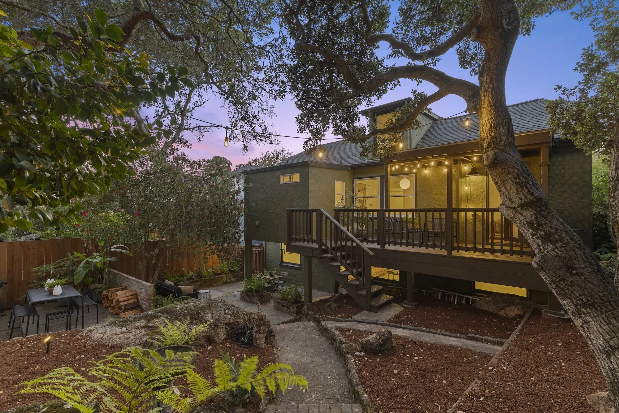 A backyard with a large tree, outdoor furniture, and a house illuminated at dusk, with string lights hanging above the deck.