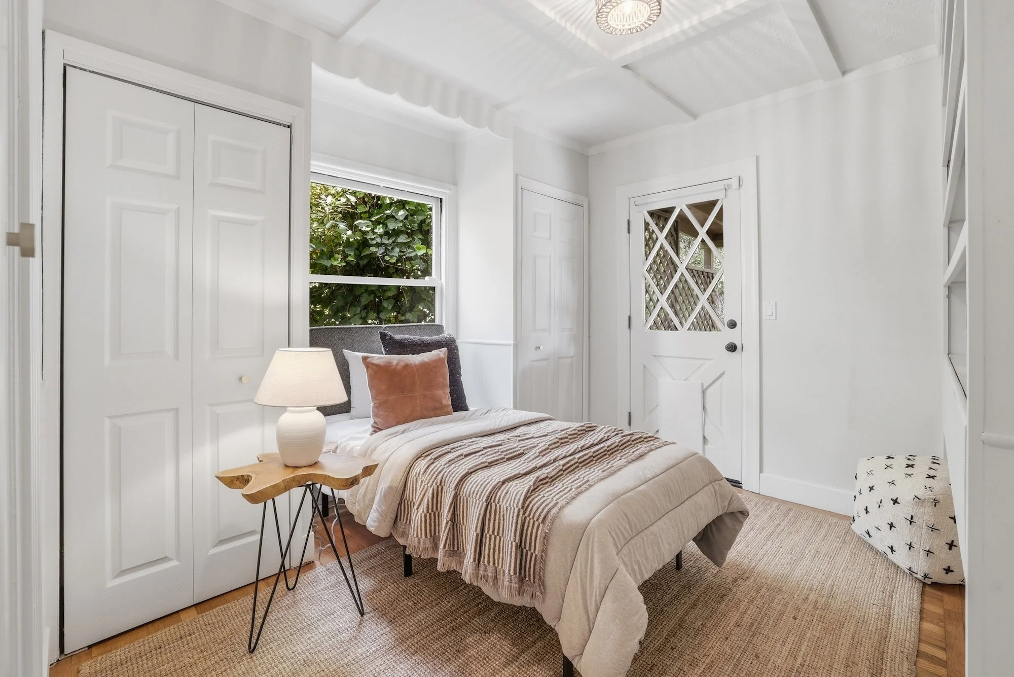 A cozy bedroom with a bed covered in beige and striped bedding, accented with pillows in shades of grey and peach. A unique wooden bedside table holds a white lamp. The room features white walls, a window showing green foliage outside, and a white do