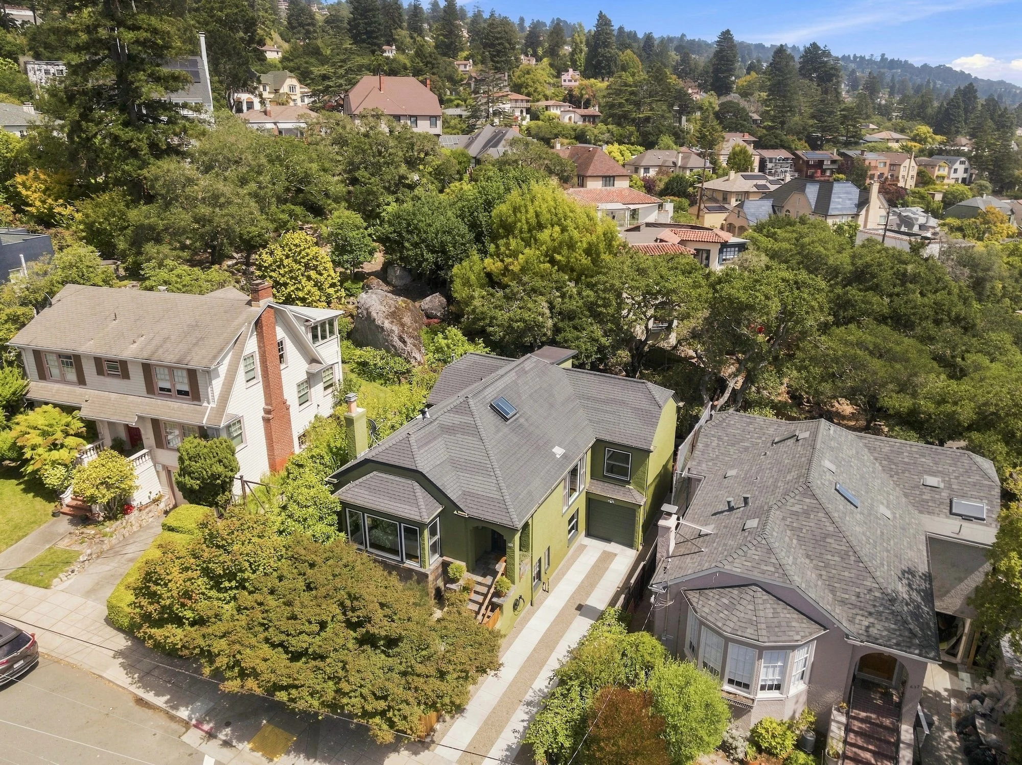 A residential neighborhood on a hillside with numerous houses surrounded by green trees, under a blue sky with some clouds.