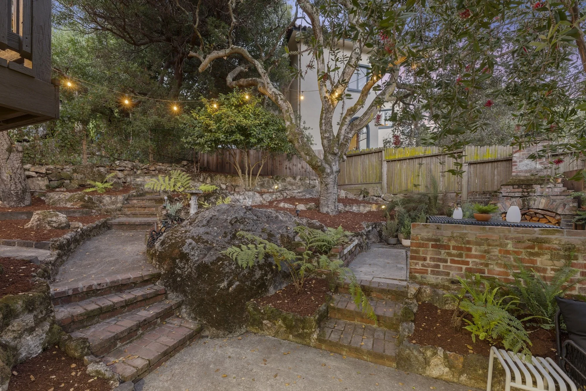 A backyard garden with a central tree, string lights, stone and brick steps, a stone garden bed, potted plants, a wooden fence, and a building in the background.