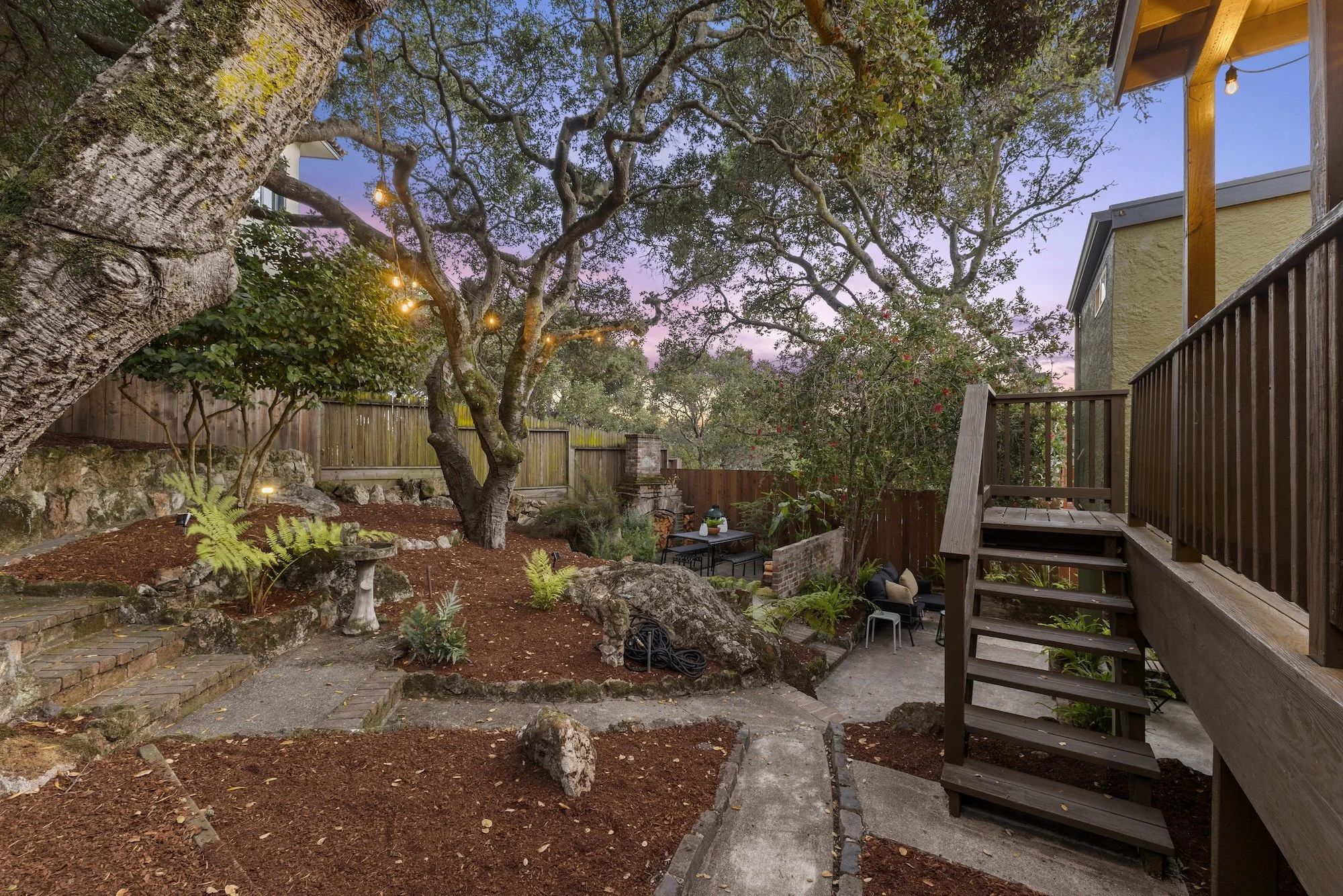 The backyard features large trees, a wooden deck, string lights, and garden furniture under a pink and blue evening sky.