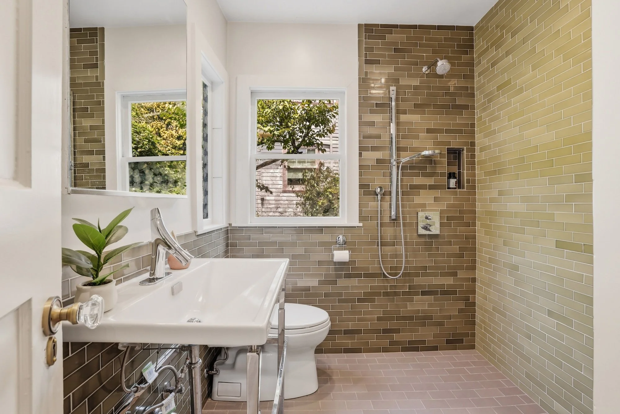 A bathroom with tan and beige brick tiles on the walls, with a walk-in shower area featuring a showerhead, a modern white toilet, and a small green plant on the sink counter. Two windows with a view of trees and buildings outside.