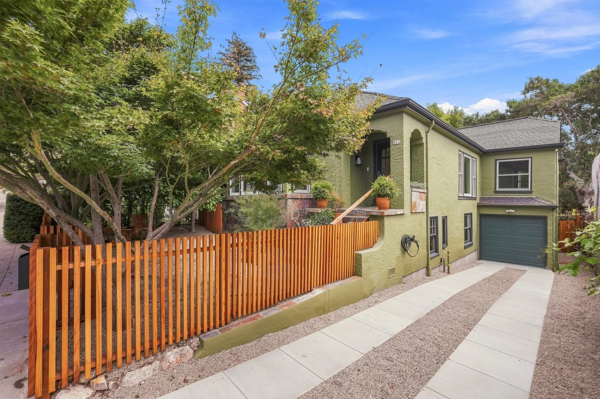 A residential house with a green exterior, gray roof, and a large front window. There is a wooden staircase with potted plants leading to the front door, and an orange wooden fence surrounds a small yard with trees and shrubbery. The driveway has con