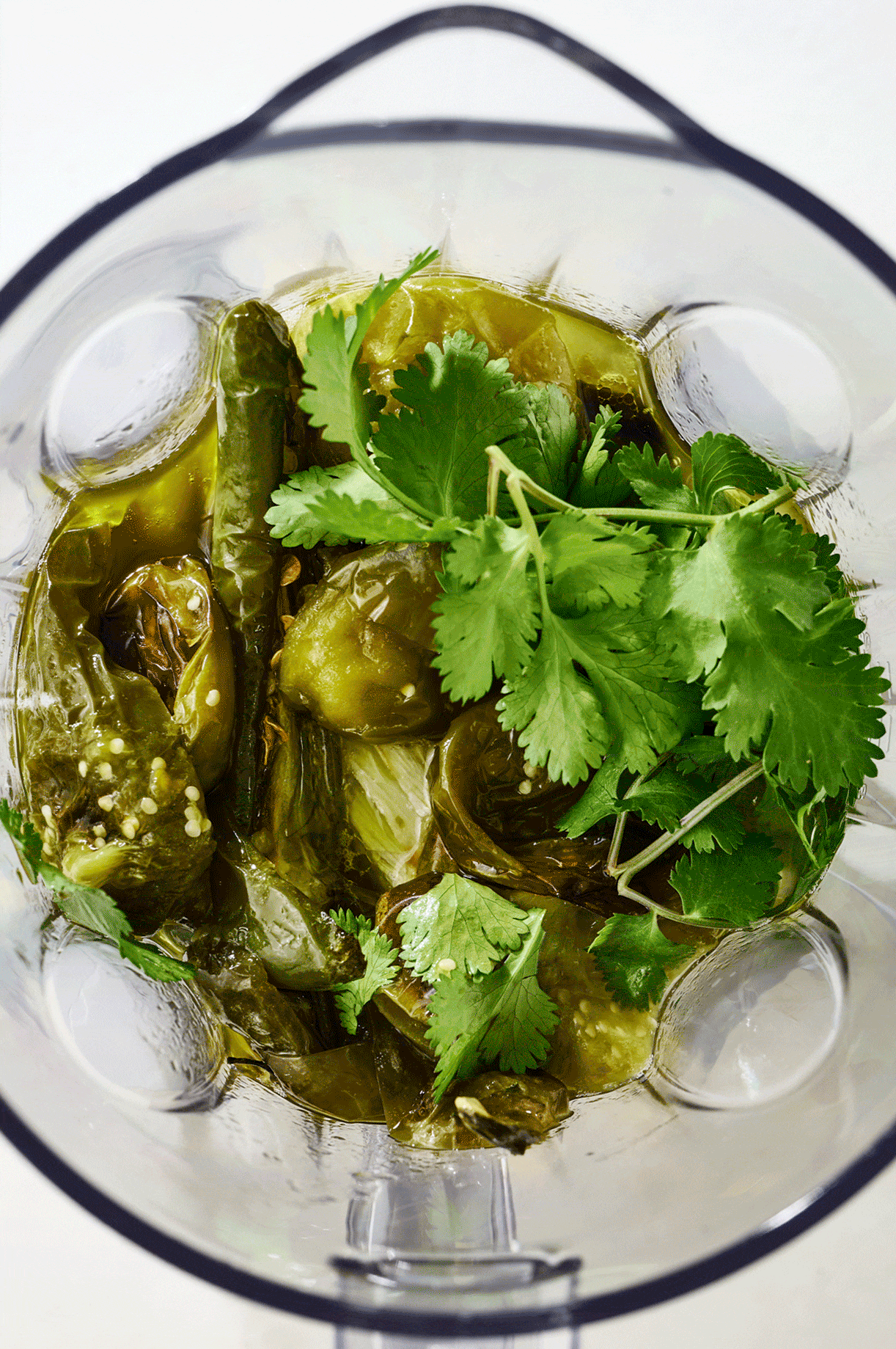 Overhead view of roasted jalapeños, tomatillos, onions and cilantro being blended to create green salsa.