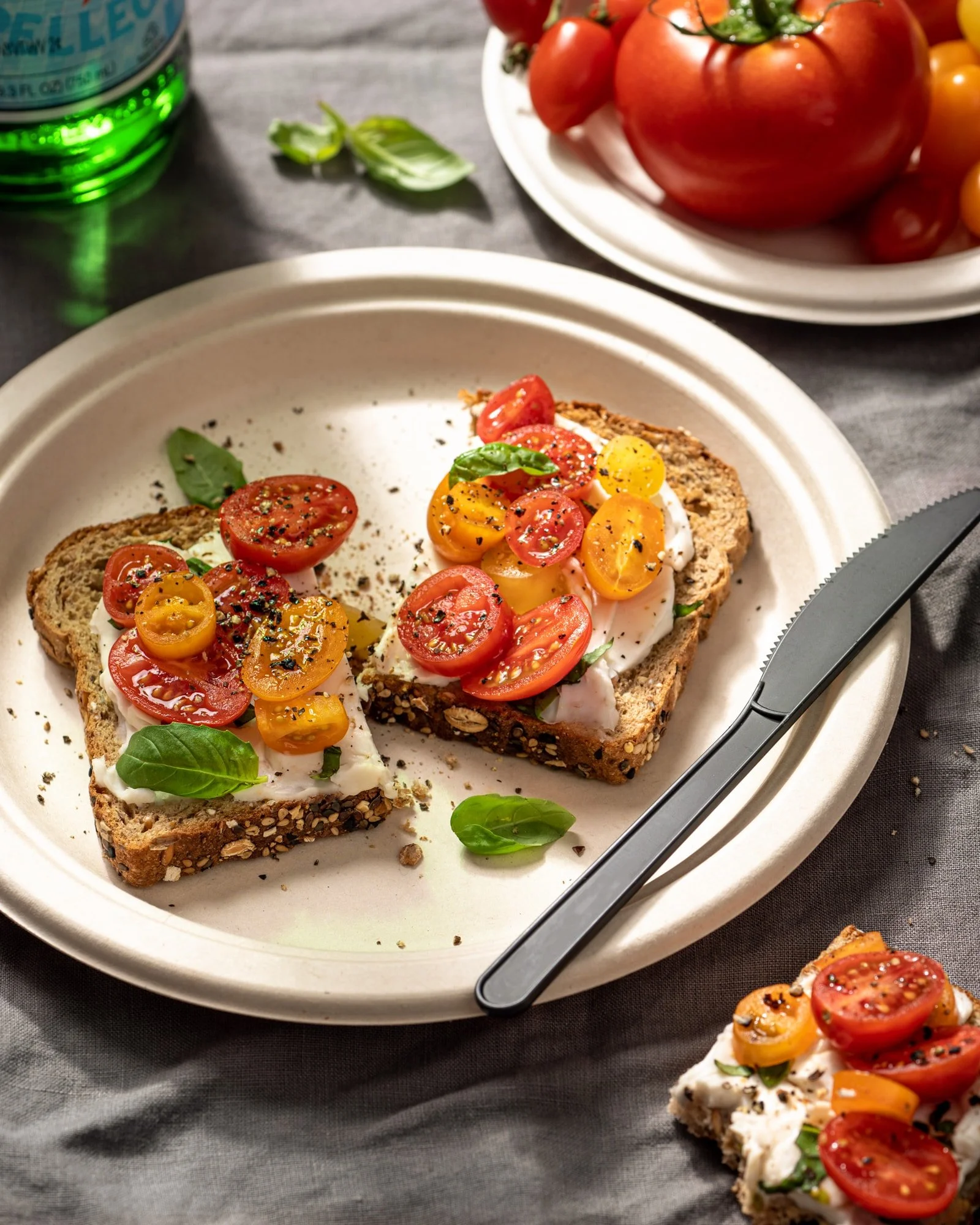 Open face toast with tomatoes, basil, and seasoning, on a white plate with a black-handled serrated knife, on a dark table with a plate of heirloom tomatoes and basil in the background.