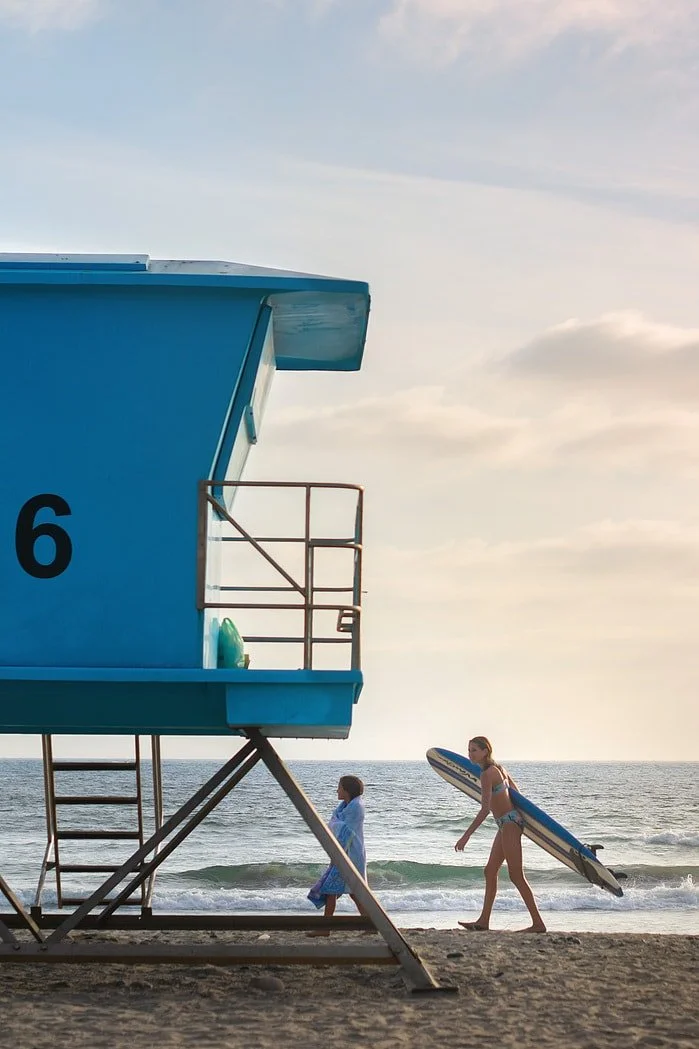 Beach scene with two kids walking near a blue lifeguard tower, one holding a surfboard, ocean waves in the background, cloudy sky.