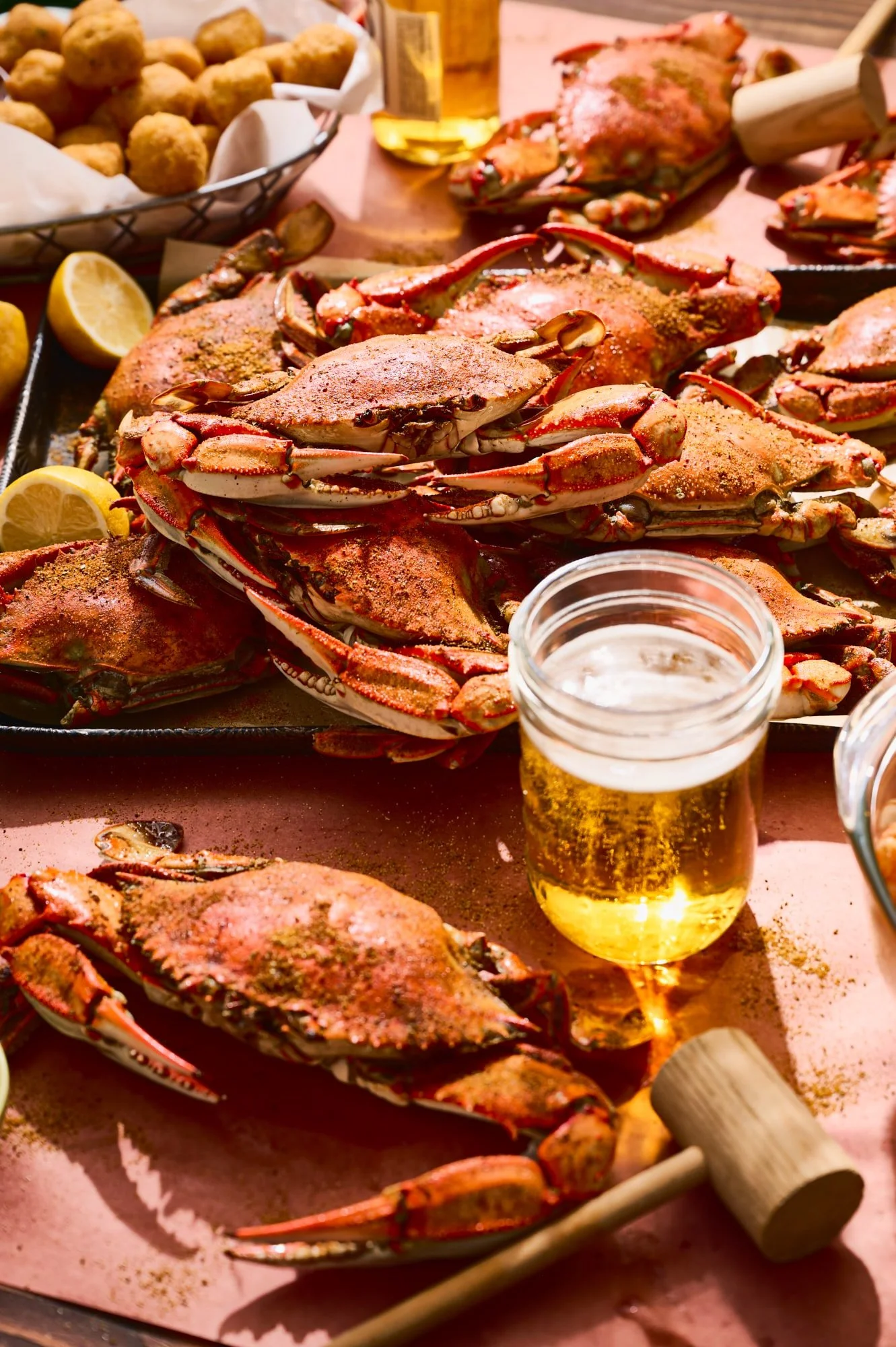 Boiled crabs seasoned with spices on a tray, with lemon wedges, beer, and wooden mallets for cracking the shells, all on a pink table.