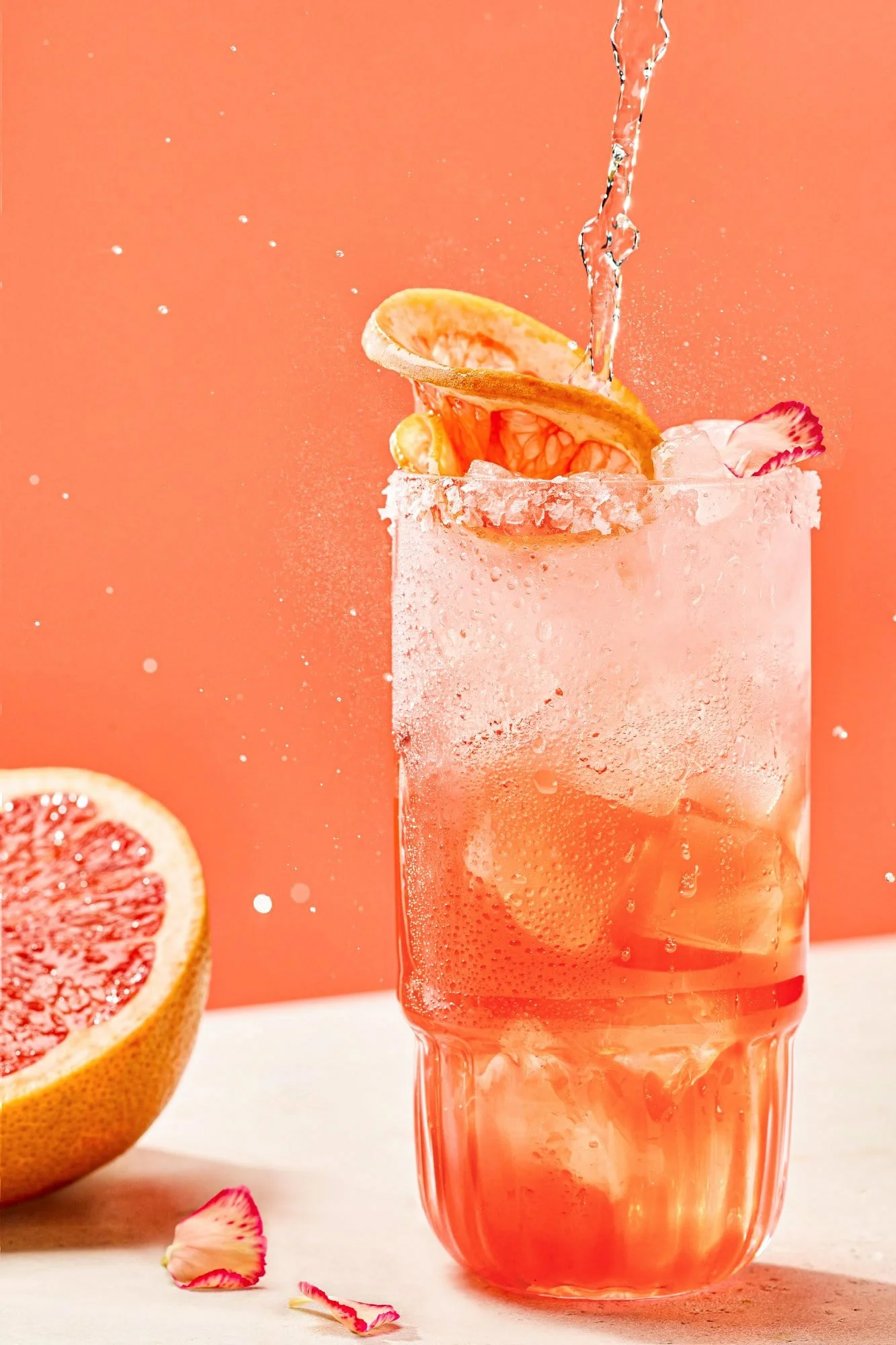 A pink grapefruit soda with ice in a tall glass, garnished with grapefruit slices and rose petal. Water is being poured into the glass, creating condensation on the outside. Whole grapefruit halves and scattered rose petals are nearby on a white surface with a pink background.