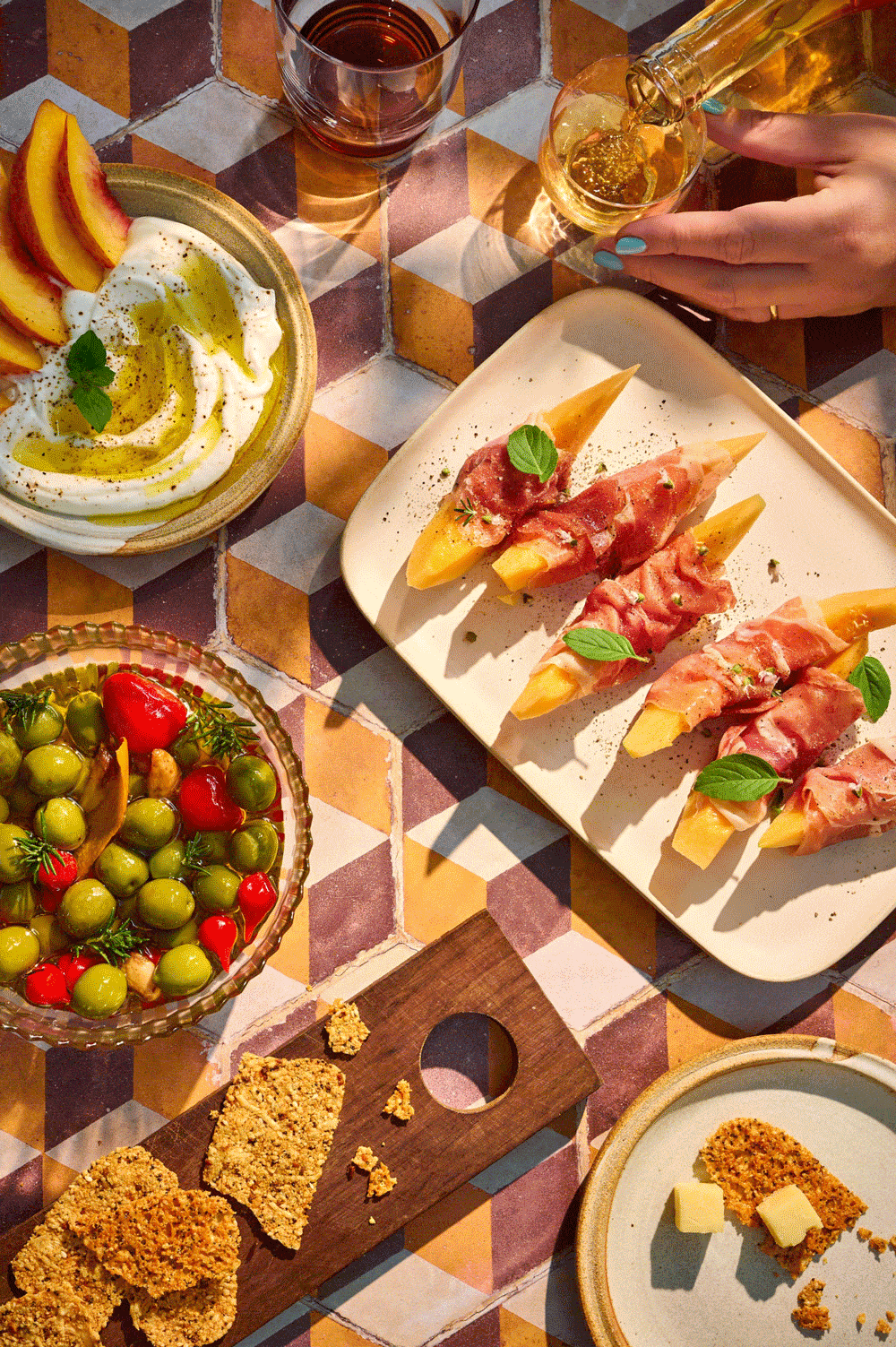 Overhead Mediterranean-inspired spread with prosciutto-wrapped melon, hummus with peaches, heirloom tomatoes, crackers, buttered bread, and drinks on a colorful geometric table.