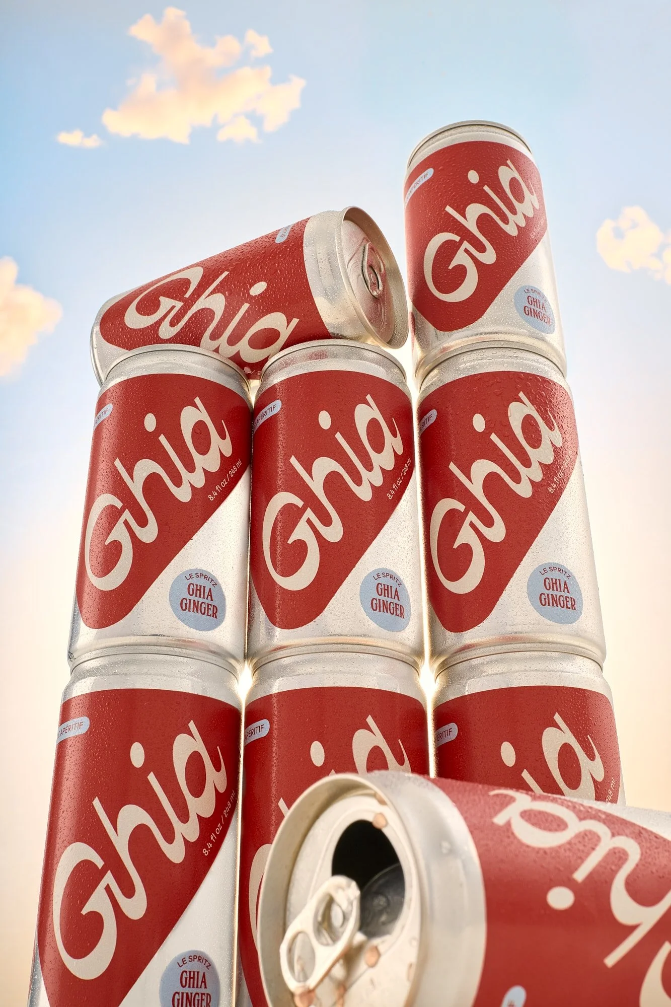 Stack of chilled red-and-white Ghia ginger sparkling beverage cans against a blue sky with clouds