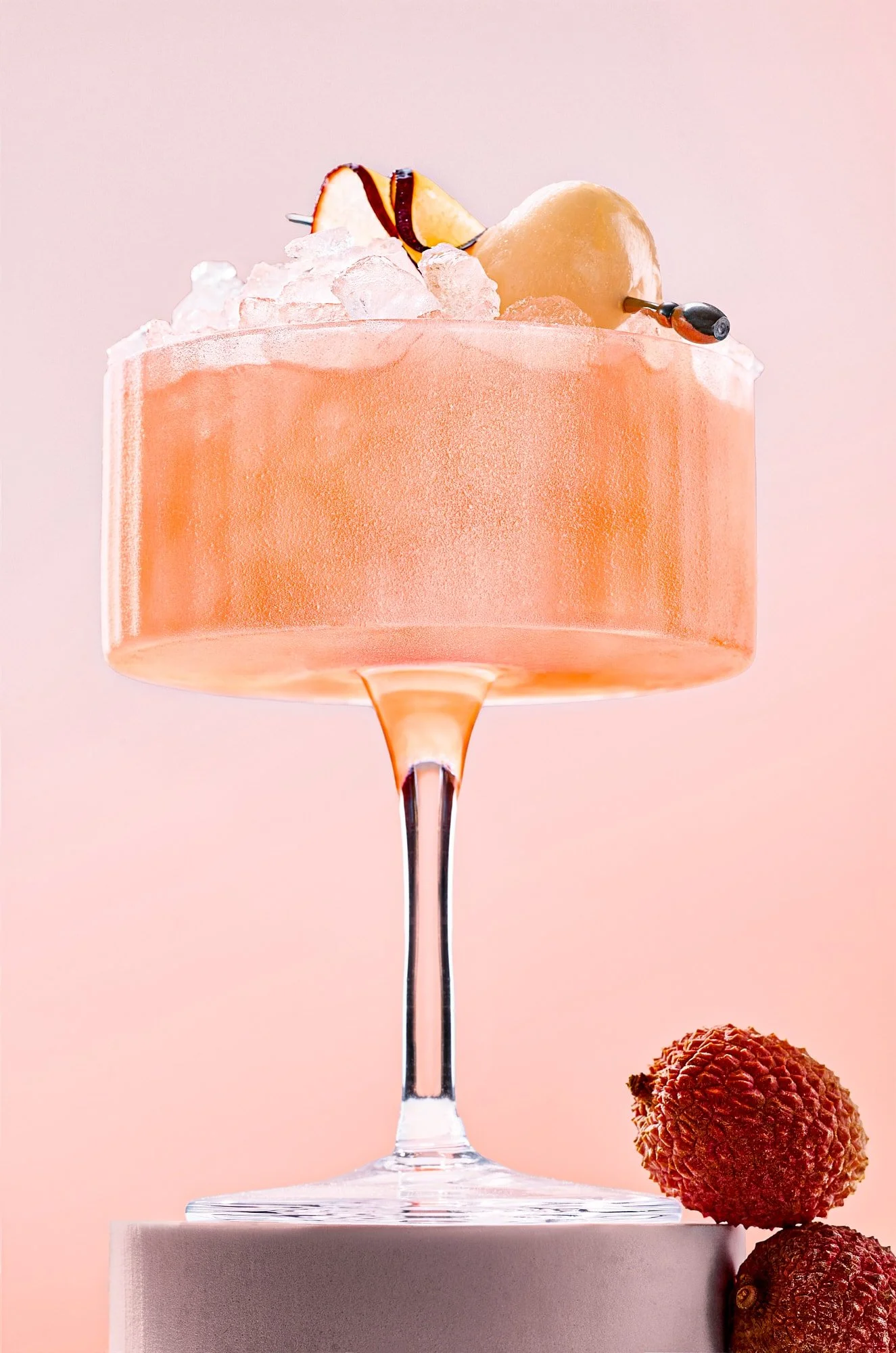 Pink cocktail with crushed ice, peach slices, and blueberries in a coupe glass, with lychees beside it