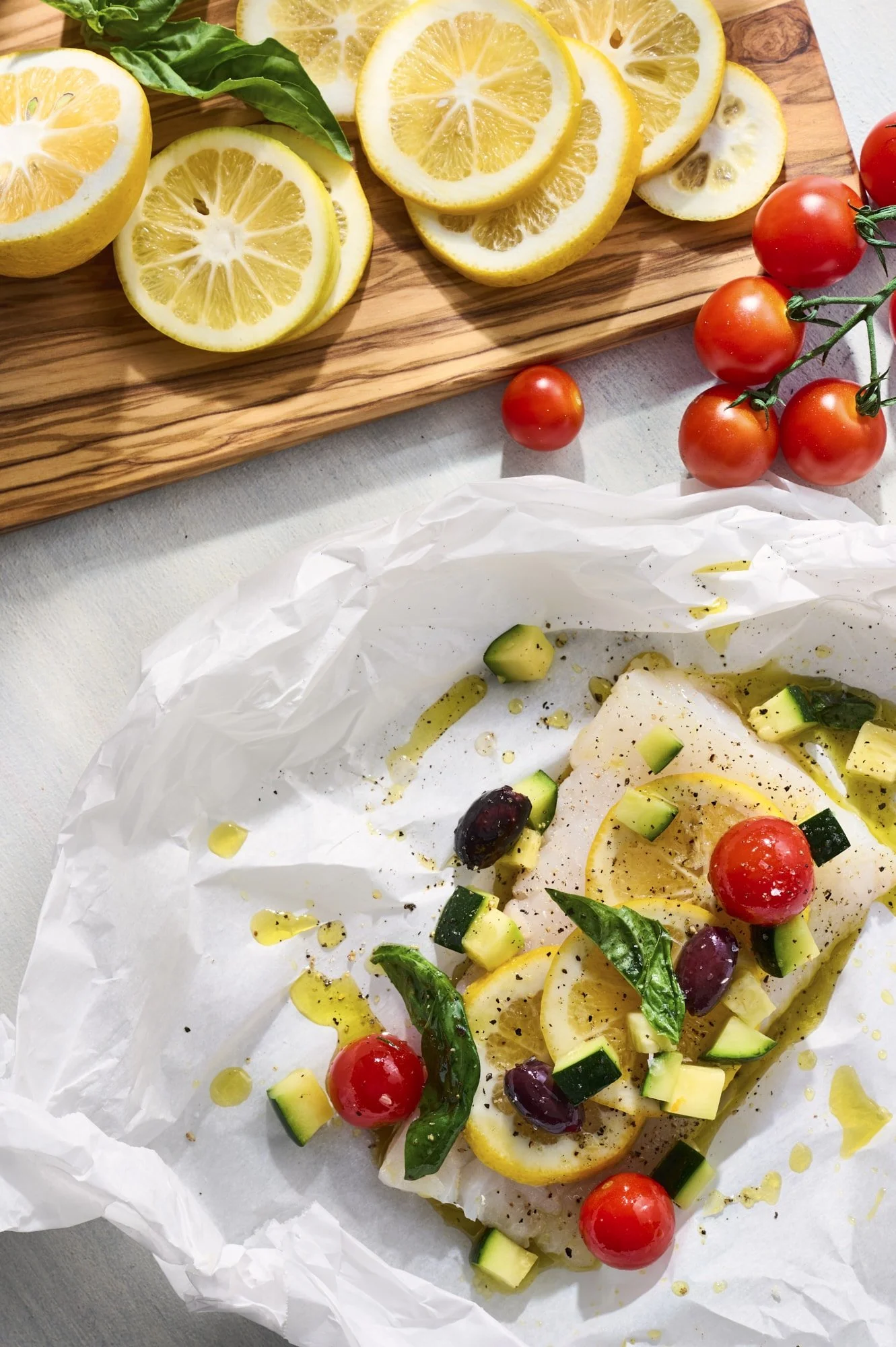 Sliced lemon and cherry tomatoes on a wooden cutting board, with a dish of fish topped with lemon slices, cherry tomatoes, chopped zucchini, basil, and olives, drizzled with olive oil.