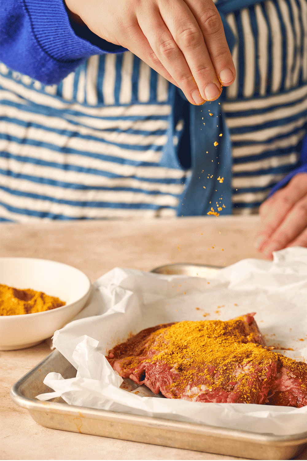 A person sprinkling spice onto a raw piece of meat on a baking sheet lined with parchment paper.