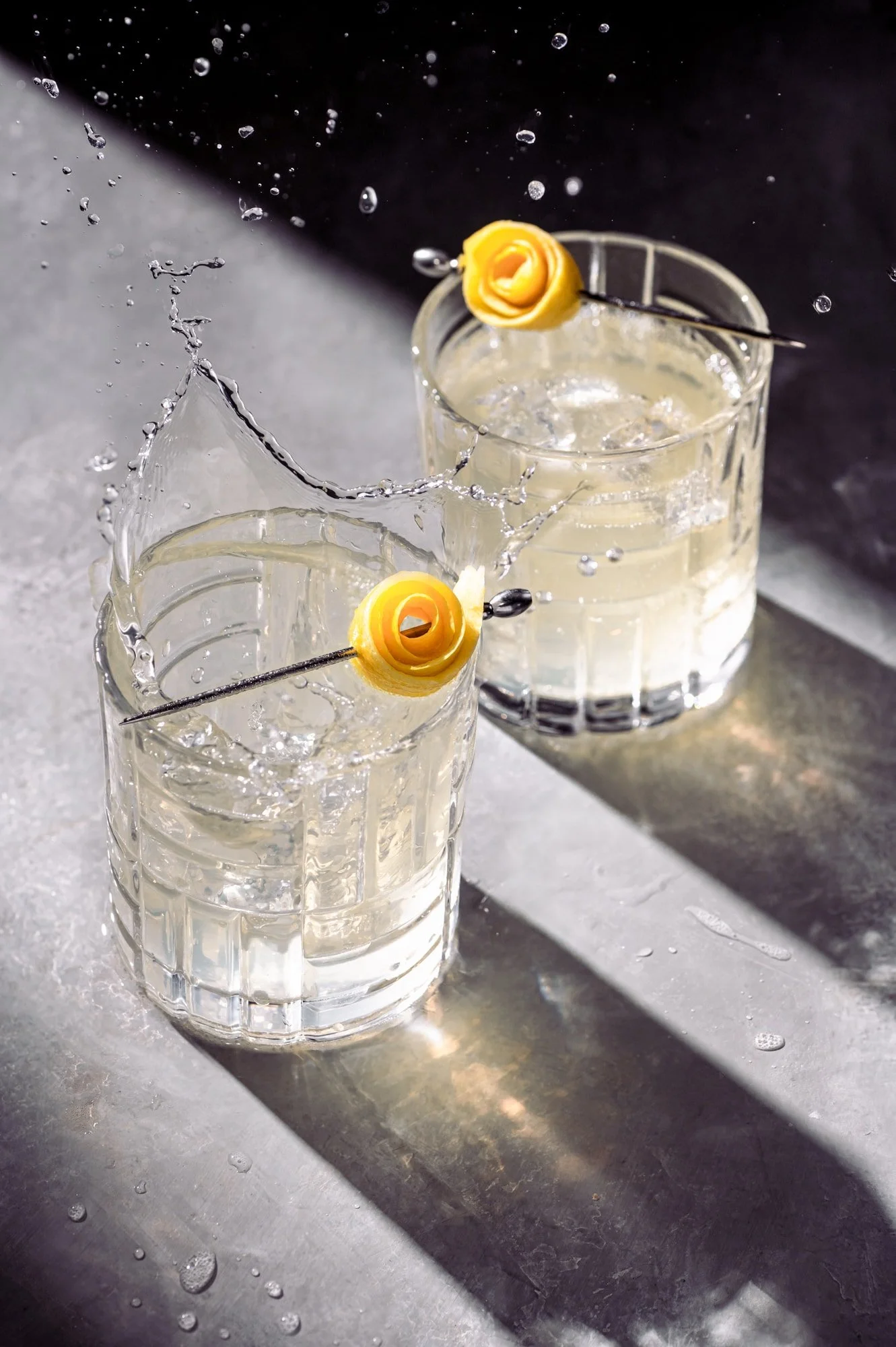 Two tall glass tumblers filled with ice and a clear beverage, garnished with rolled yellow cheese slices on metal skewers, placed on a wet, reflective metallic surface.