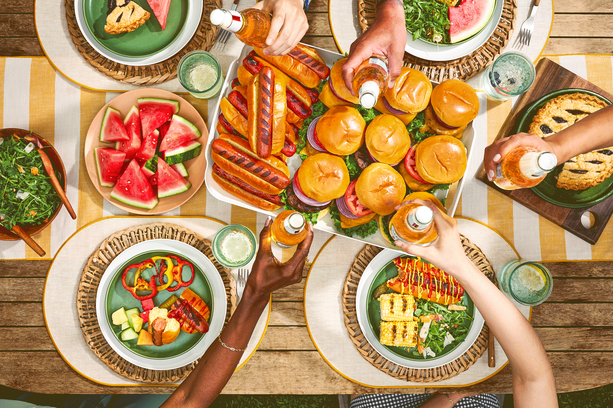 Overhead view of a diverse group enjoying an outdoor summer barbecue, clinking Michelob Ultras over a table filled with sliders, hot dogs, watermelon, salad, and grilled vegetables.
