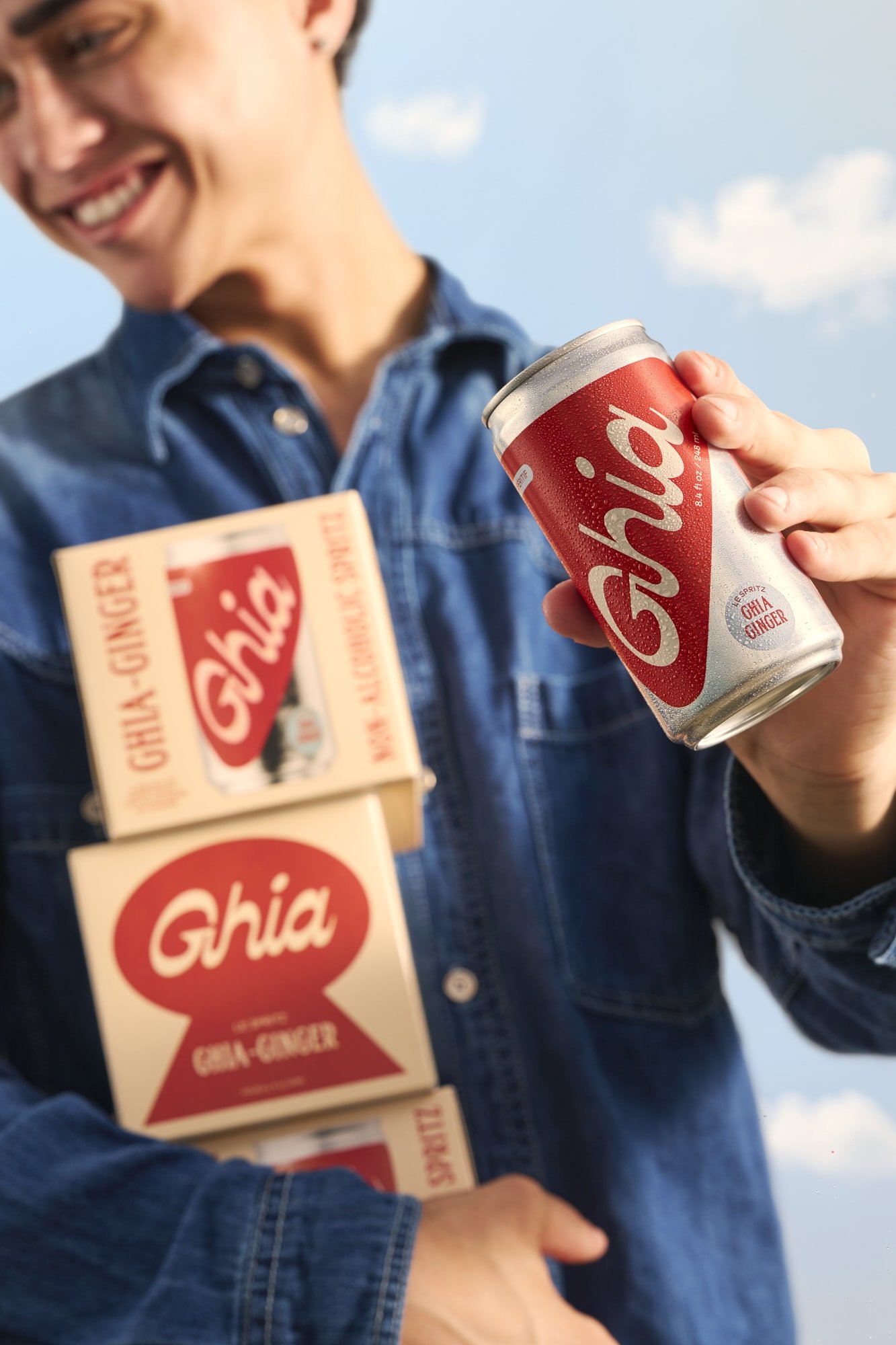 Smiling man in a denim shirt holding a chilled Ghia ginger spritz can and stacked Ghia Ginger beverage boxes