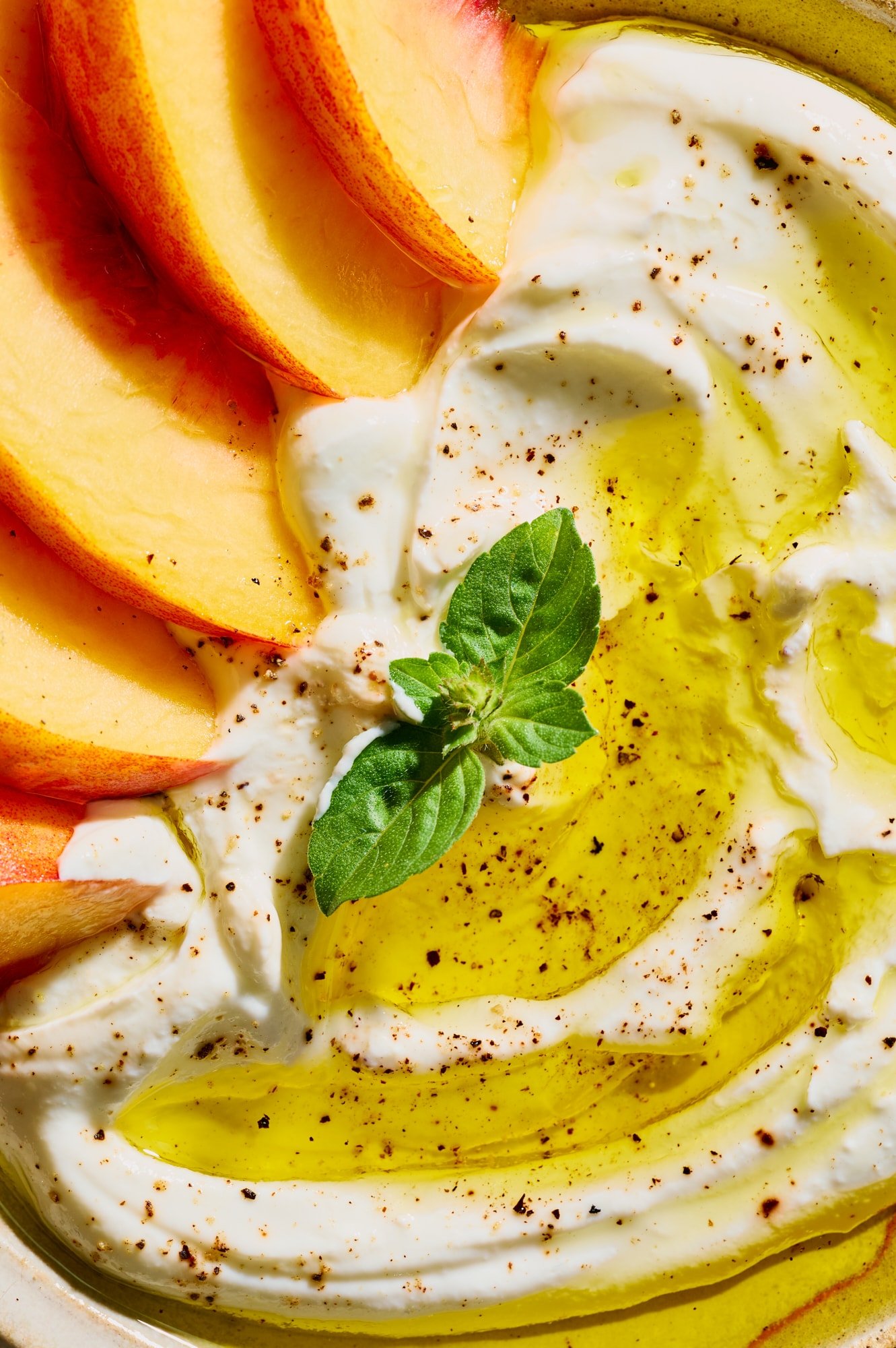 Close-up of Greek yogurt topped with peach slices, olive oil, black pepper, and fresh basil.