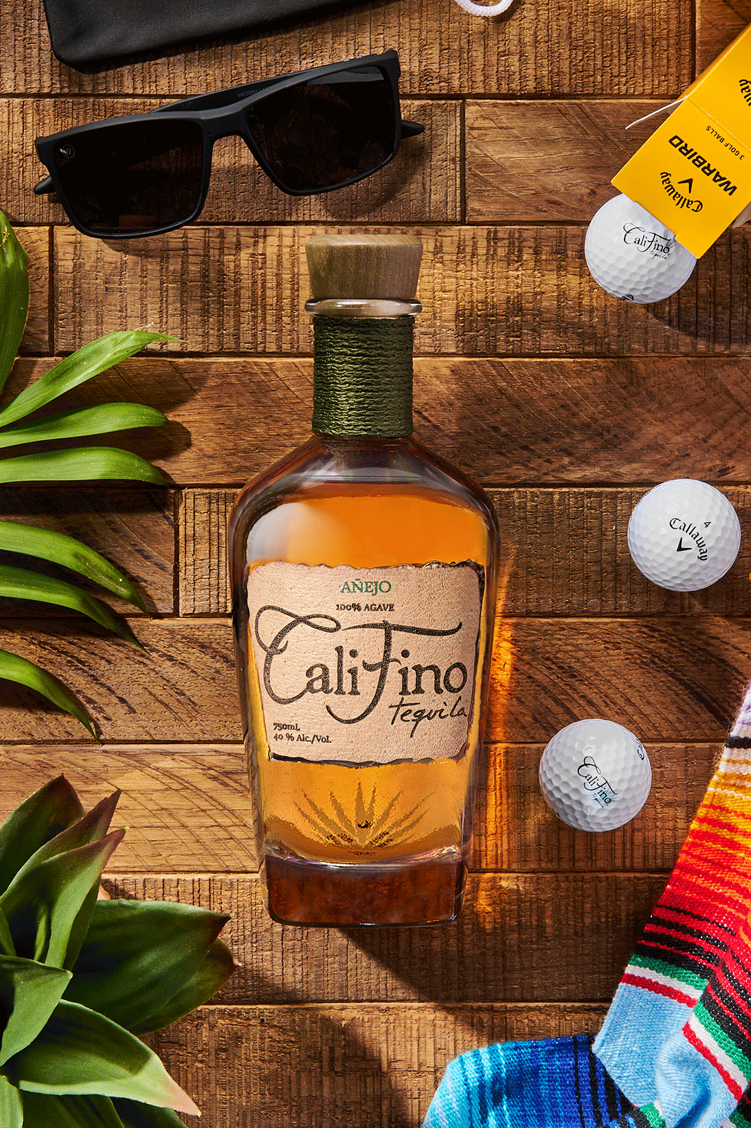 Califino Añejo tequila bottle surrounded by golf balls, sunglasses, agave leaves, and a colorful woven blanket on a wooden table