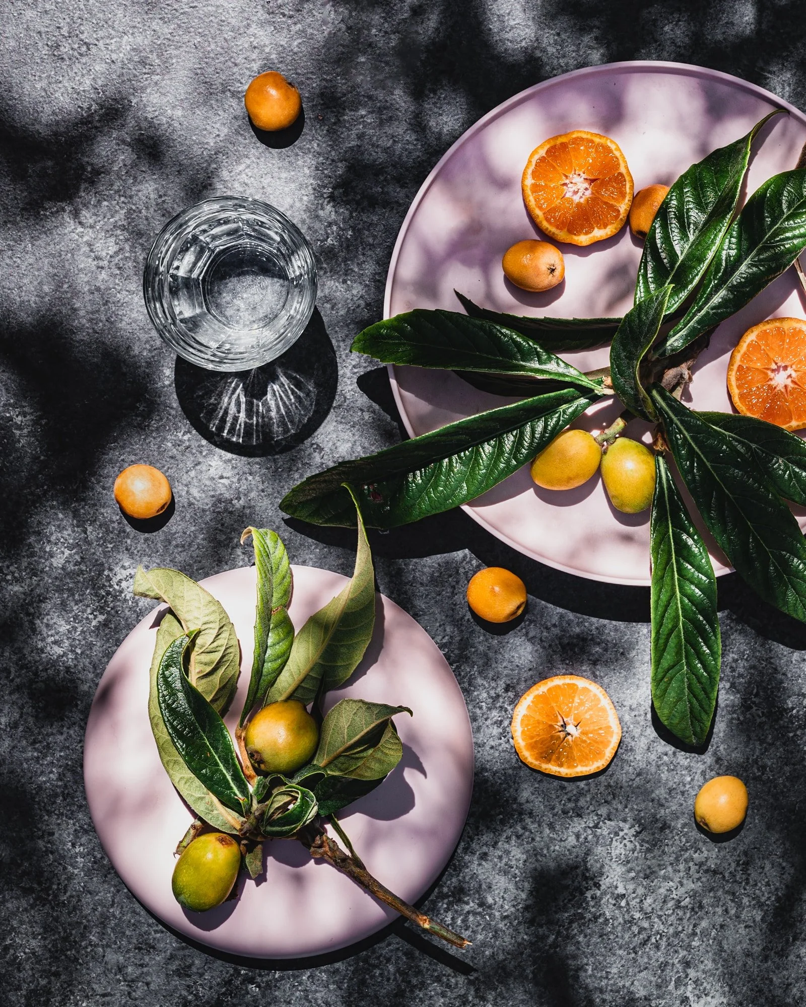 A dark gray textured surface with a pink plate holding a branch with green leaves and green fruits, halved oranges, and a few scattered oranges and fruits around the plate, along with a clear drinking glass filled with water.