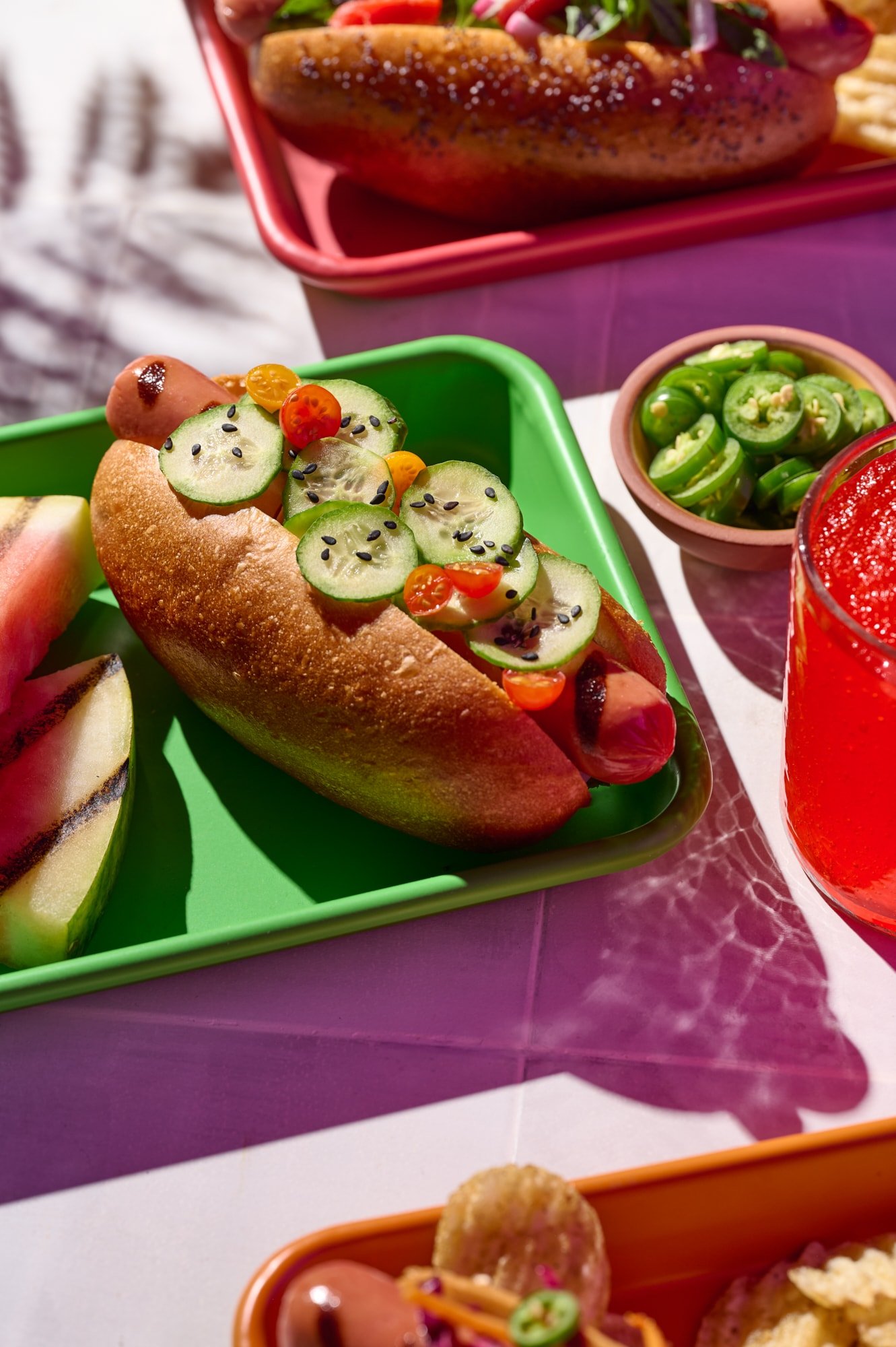 Hot dog topped with sliced cucumbers, cherry tomatoes, and black sesame seeds on a green tray, with sliced jalapeños and a red drink nearby