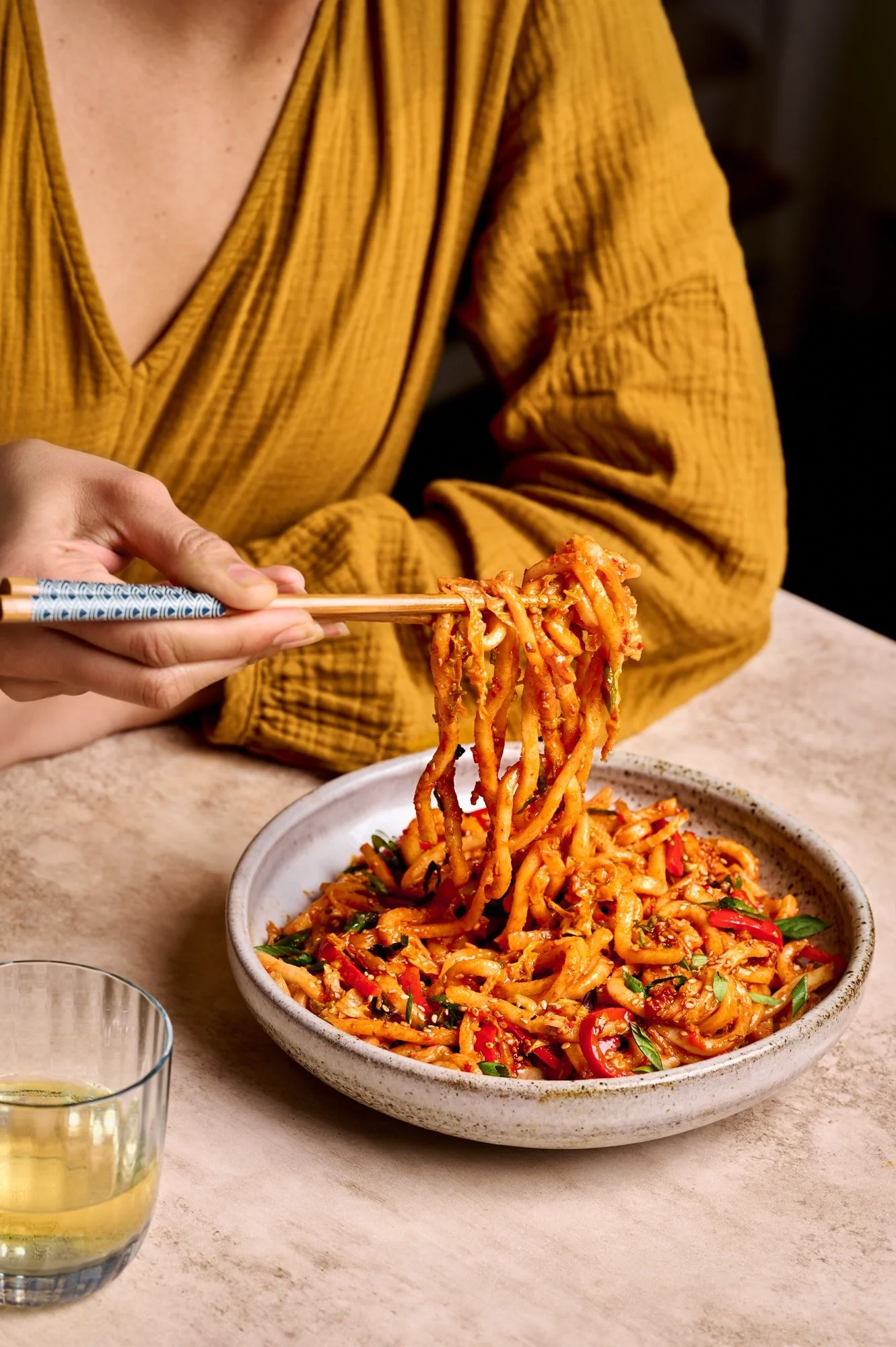 A person wearing a mustard yellow shirt uses chopsticks to lift Rainbow Plant Life’s gochujang noodles from a bowl with vegetables on a beige table, with a glass drink nearby.