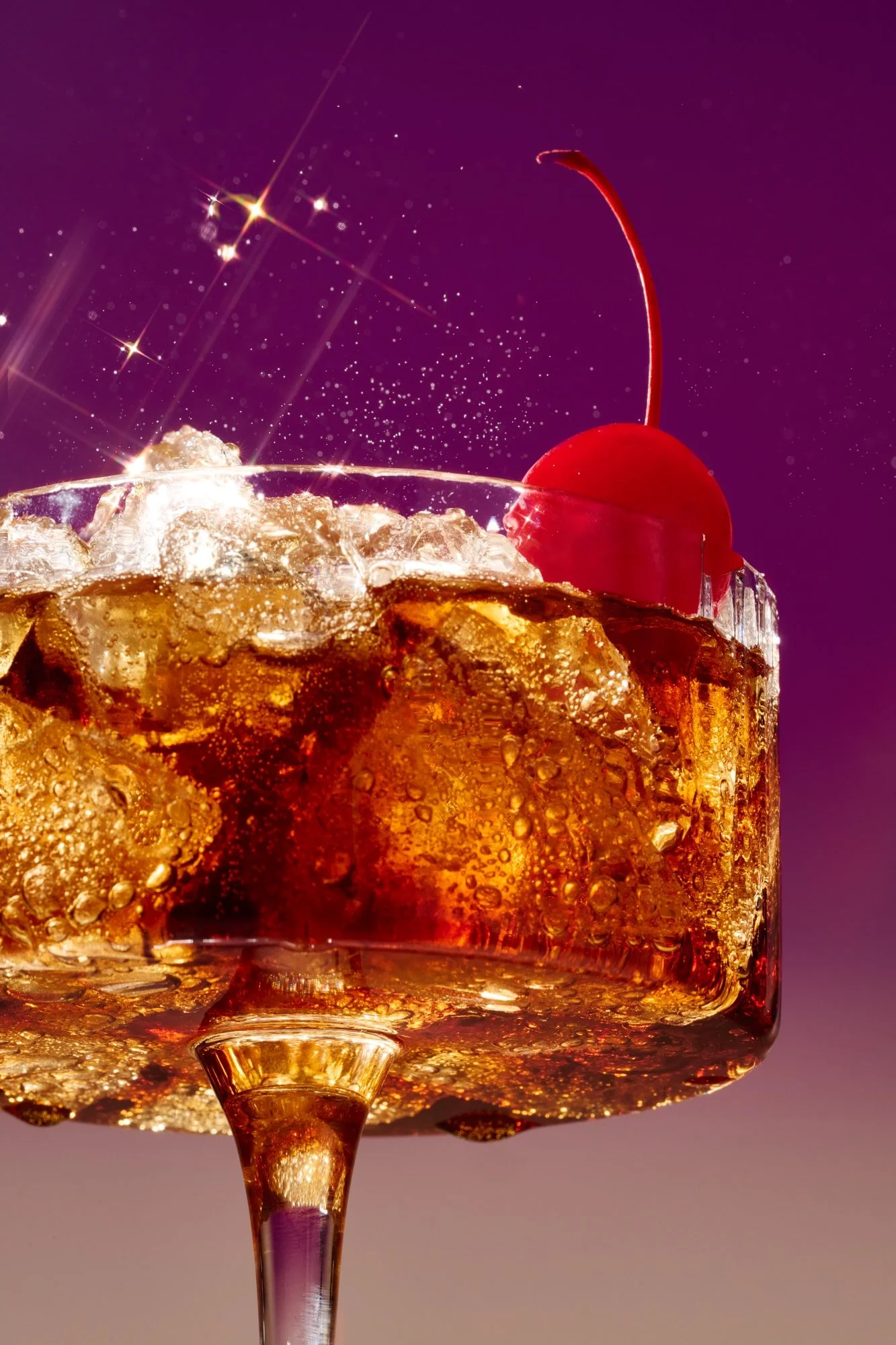 Close-up of a cherry cola cocktail with ice in a glass, garnished with a red cherry against a purple background