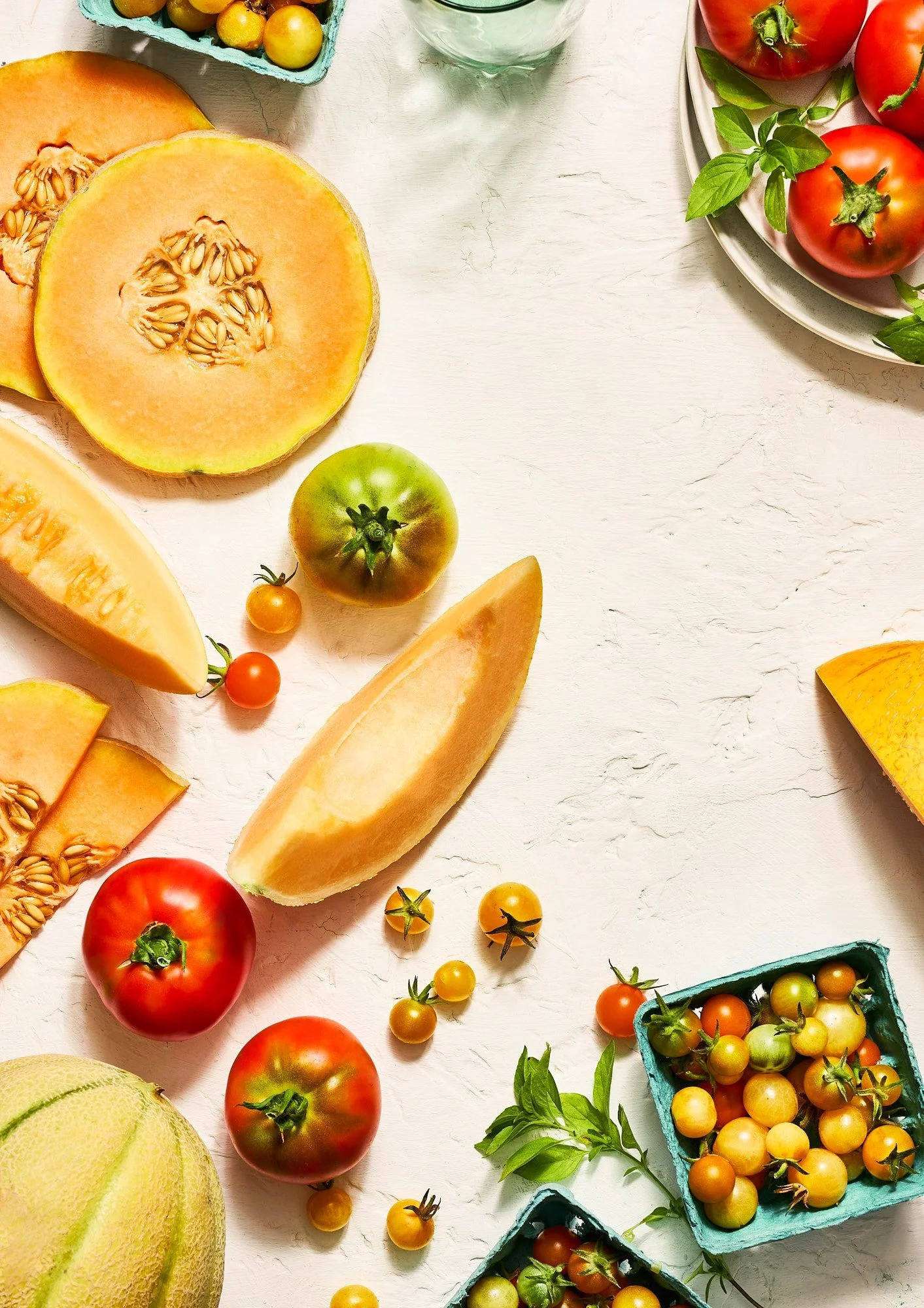 Overhead arrangement of sliced cantaloupe, tomatoes, cherry tomatoes, basil, and other melons on a white tabletop