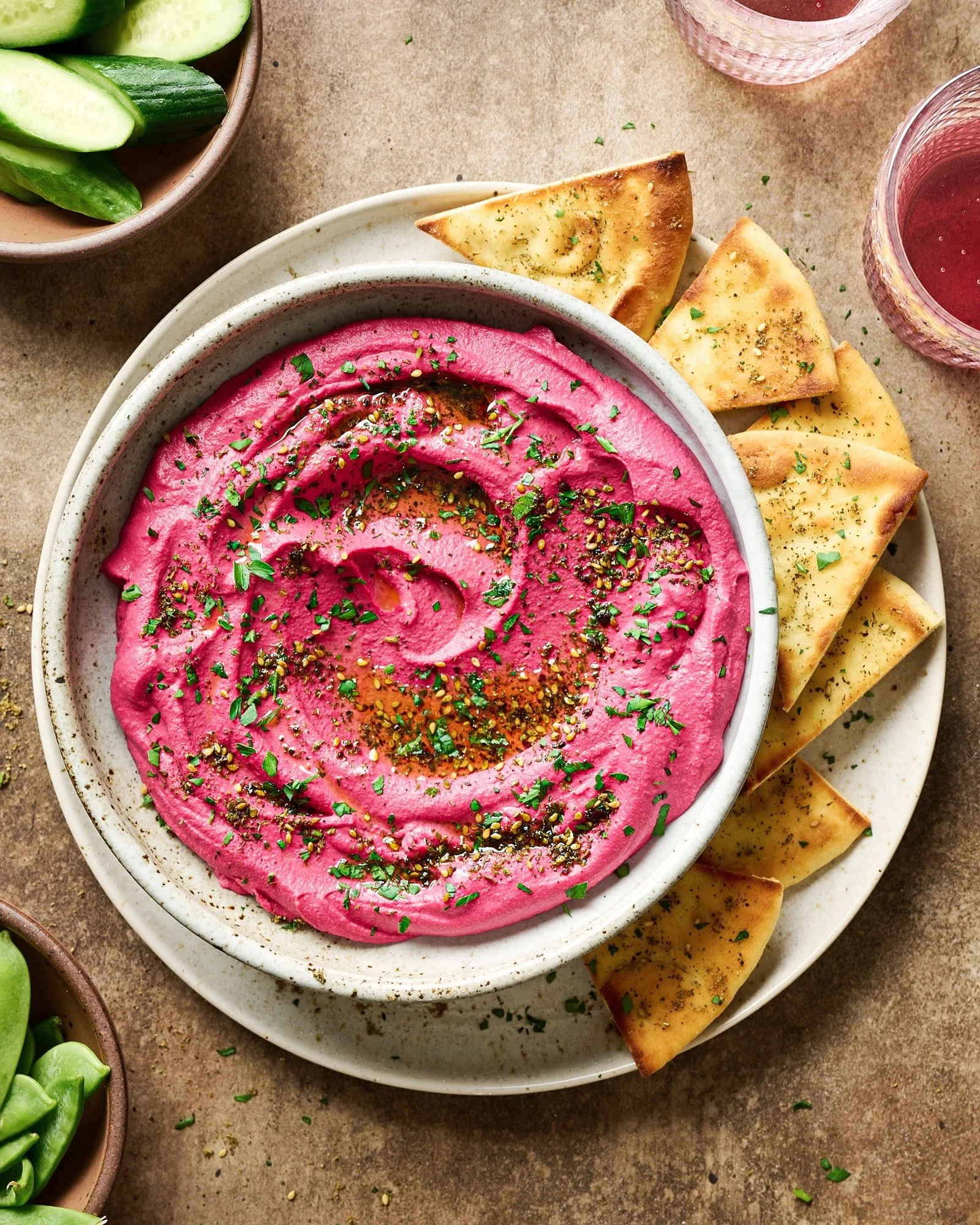 Bowl of bright pink beet hummus topped with herbs and spices, served with slices of toasted pita bread. In the background, there are glasses with pink drinks and a bowl of sliced cucumbers.