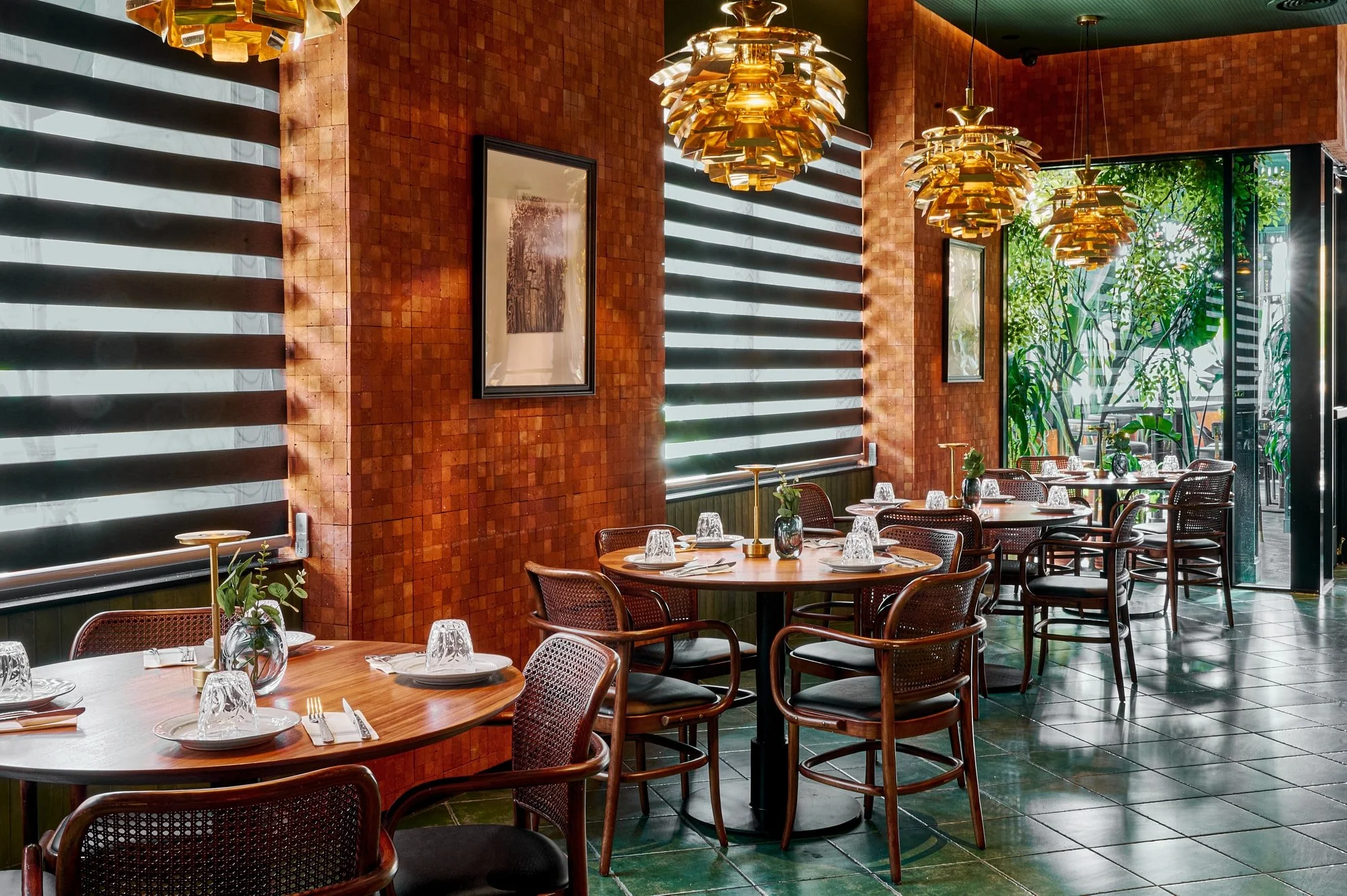 Interior of Starleaf, a Pasadena-based restaurant with wooden tables set for dining. The restaurant features warm, reddish brick walls, modern hanging gold light fixtures, and large windows with blinds allowing natural light and nature in.