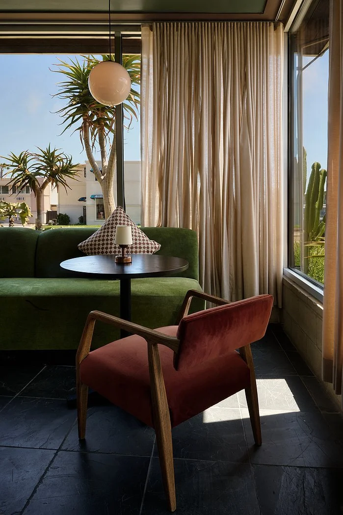Indoor seating area with a green sofa, a brown wooden armchair with reddish upholstery, a black round table, a small lamp on the table, a patterned pillow on the sofa, large windows with beige curtains, and plants outside.