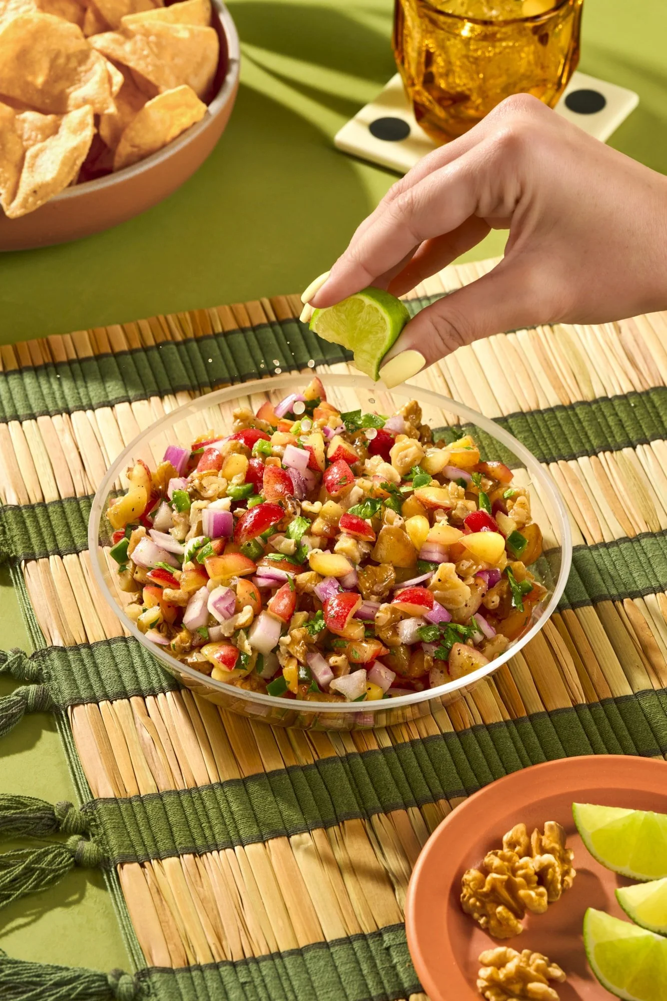 A bowl of chopped vegetables and nuts, with a hand squeezing lime over the salad. In the background, there is a bowl of tortilla chips, a glass of amber-colored beverage, and a small plate with walnuts and lime wedges. The setting appears to be a colorful, tropical-themed table.